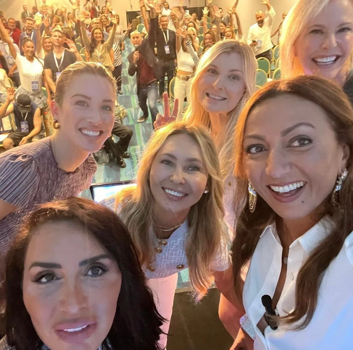 Group of women taking a selfie at a conference or event, with many people seated and standing behind them, some making peace signs, in a large, well-lit room.