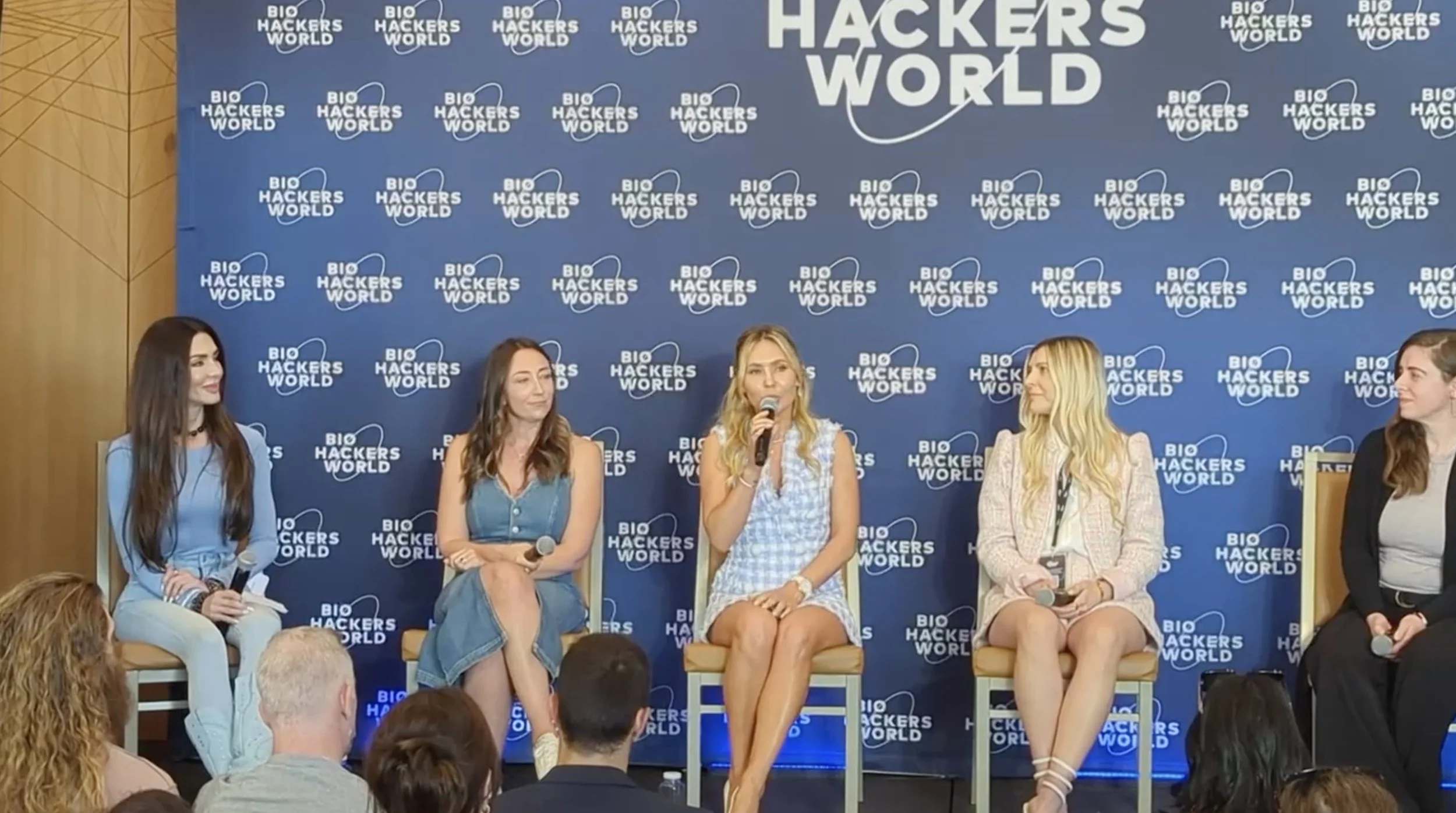 Five women sitting on chairs on stage at a panel discussion with a blue backdrop featuring the 'Bio Hackers World' logo. One woman is speaking into a microphone, while others hold microphones.