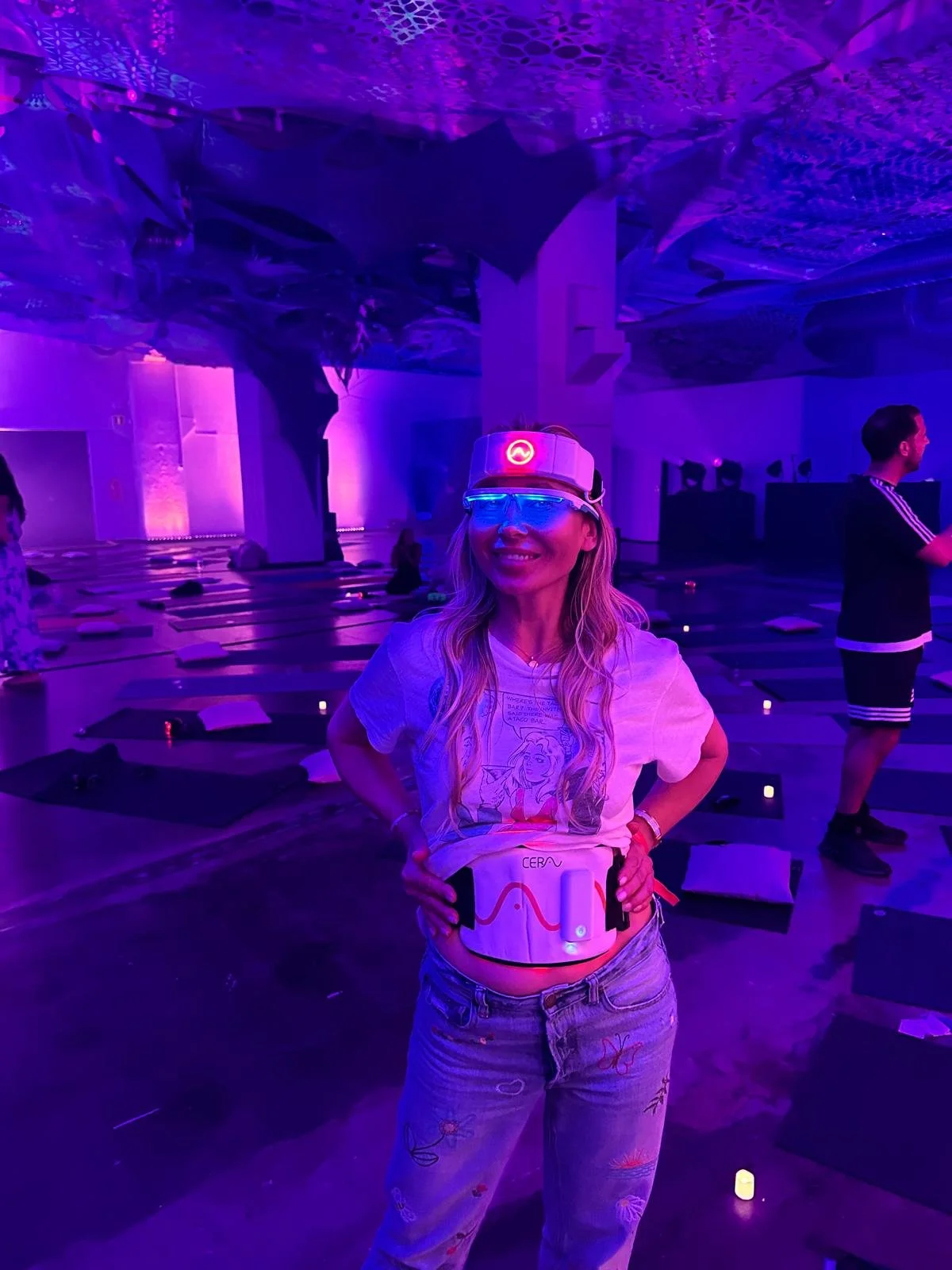 A woman wearing a headlamp and virtual reality goggles in a dark, purple-lit room with yoga mats on the floor, some people sitting in the background.