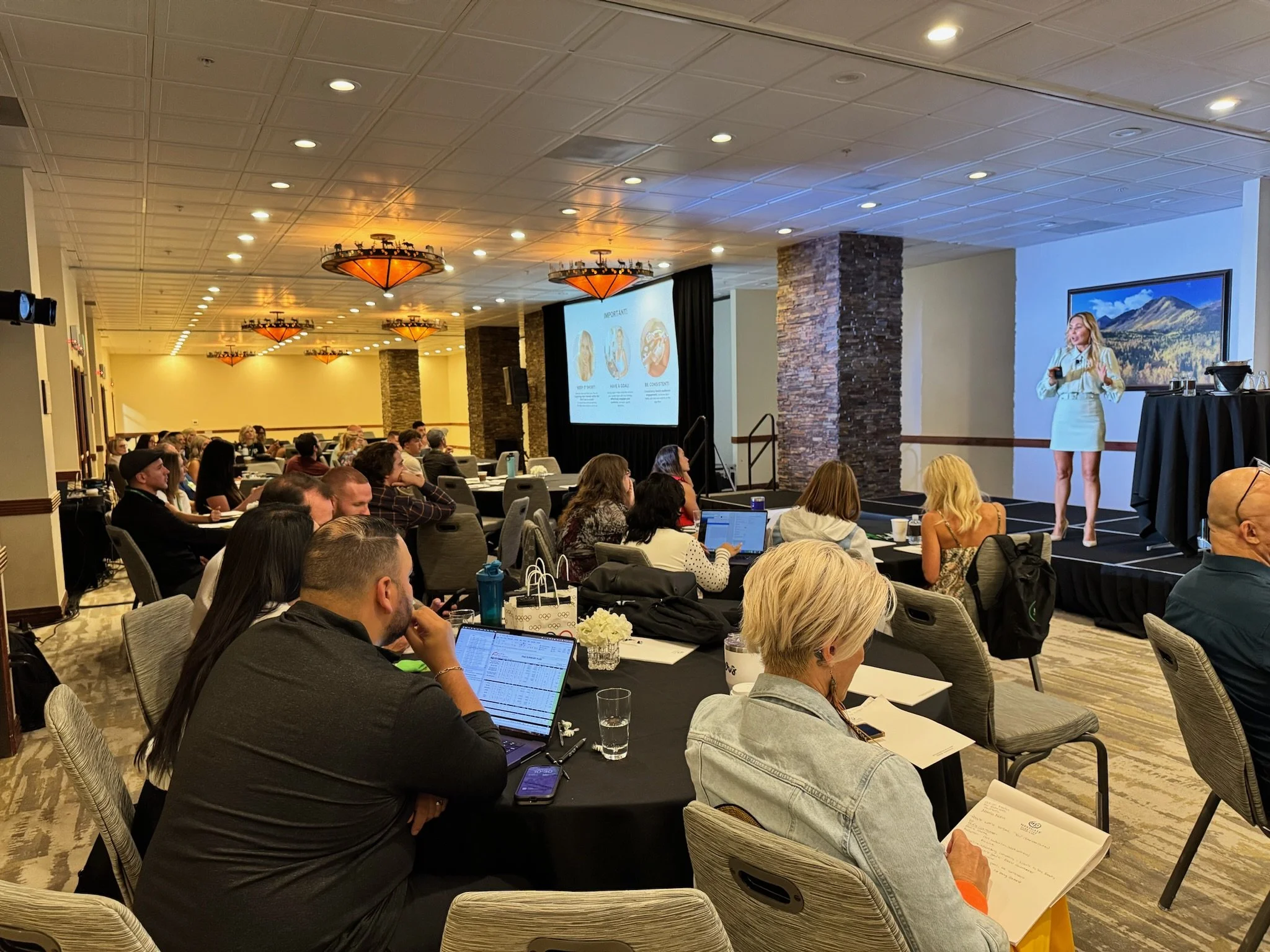 A woman in a white dress is giving a presentation on stage at a conference room filled with seated attendees. The room has chandeliers and large screens displaying her slides.