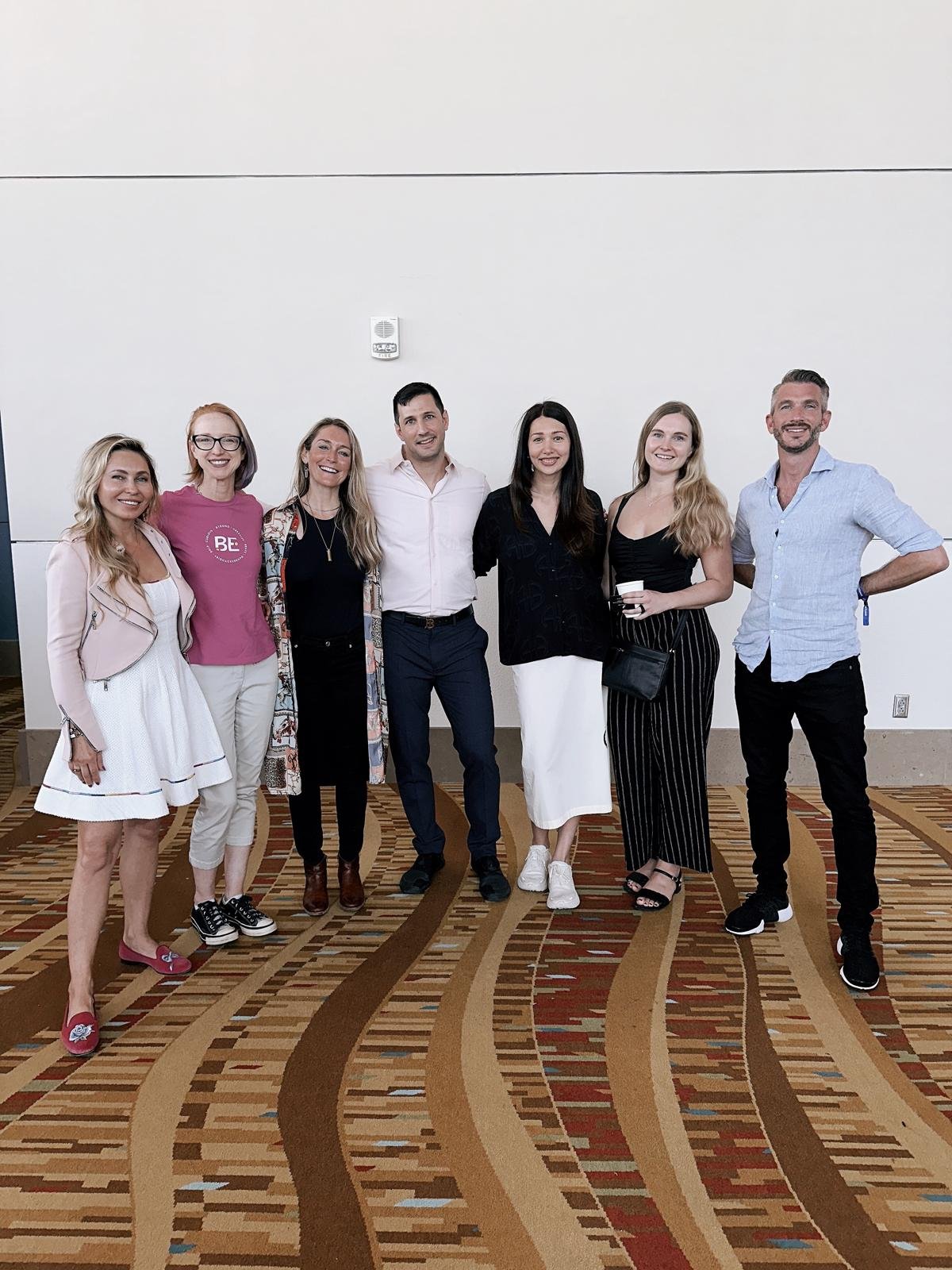 Group of seven people standing together in a room with patterned carpet and plain white wall, smiling and posing for the photo.