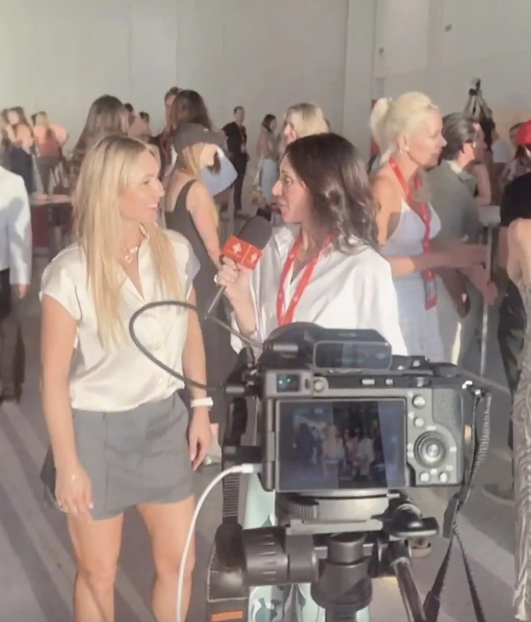 A woman being interviewed on camera at a crowded event, with multiple people in the background.
