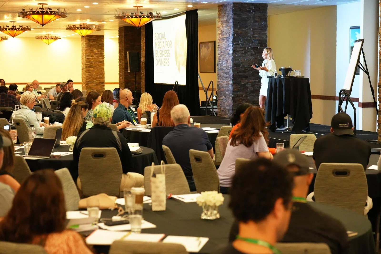 A conference room filled with attendees seated at round tables, listening to a presenter on stage with a large screen displaying a presentation titled 'Social Media for Business Owners'. The room has warm lighting and stone columns.