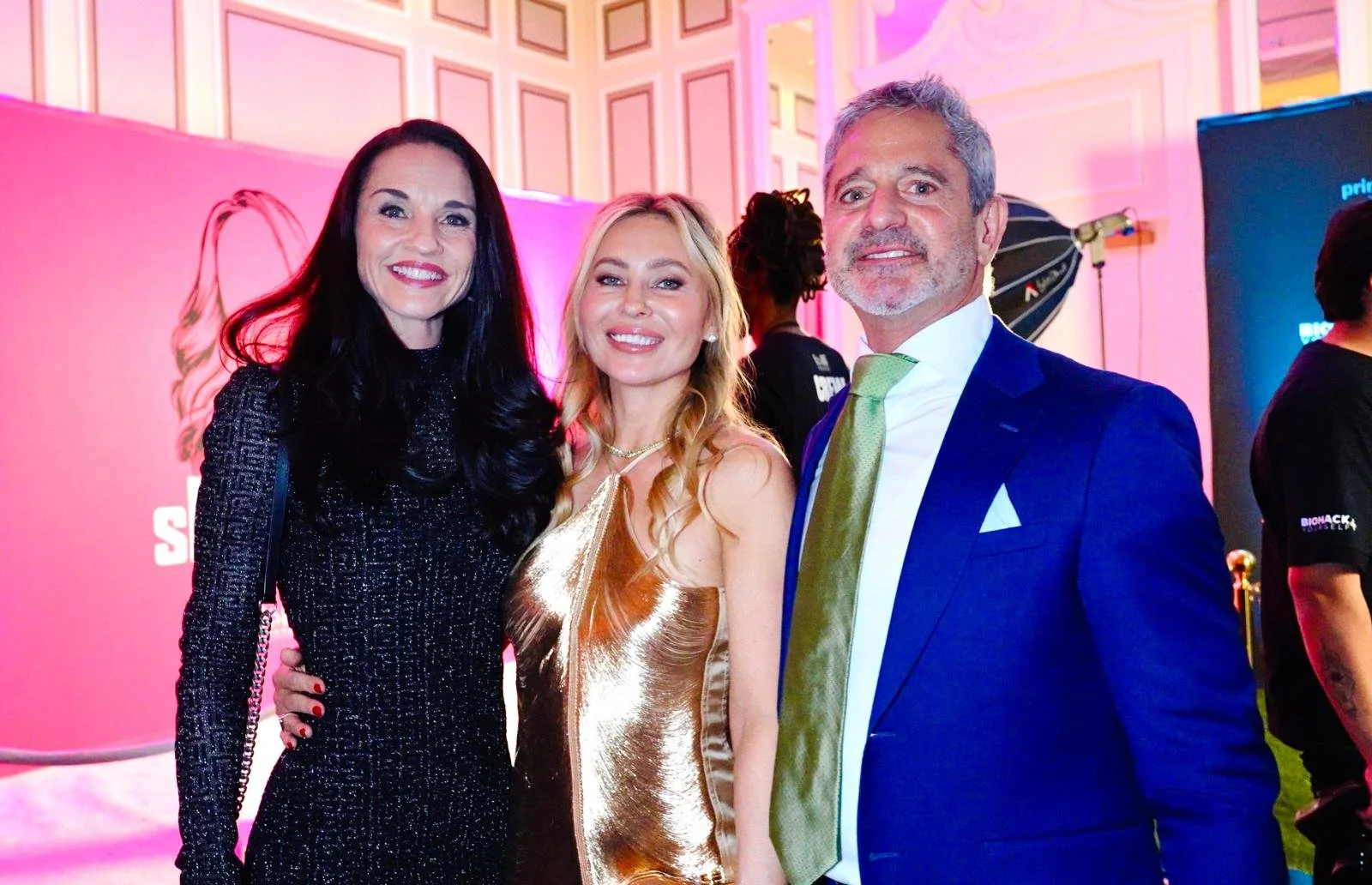 Three people smiling and posing for a photo at an event in a brightly colored, fancy interior with pink lighting and decorative walls.