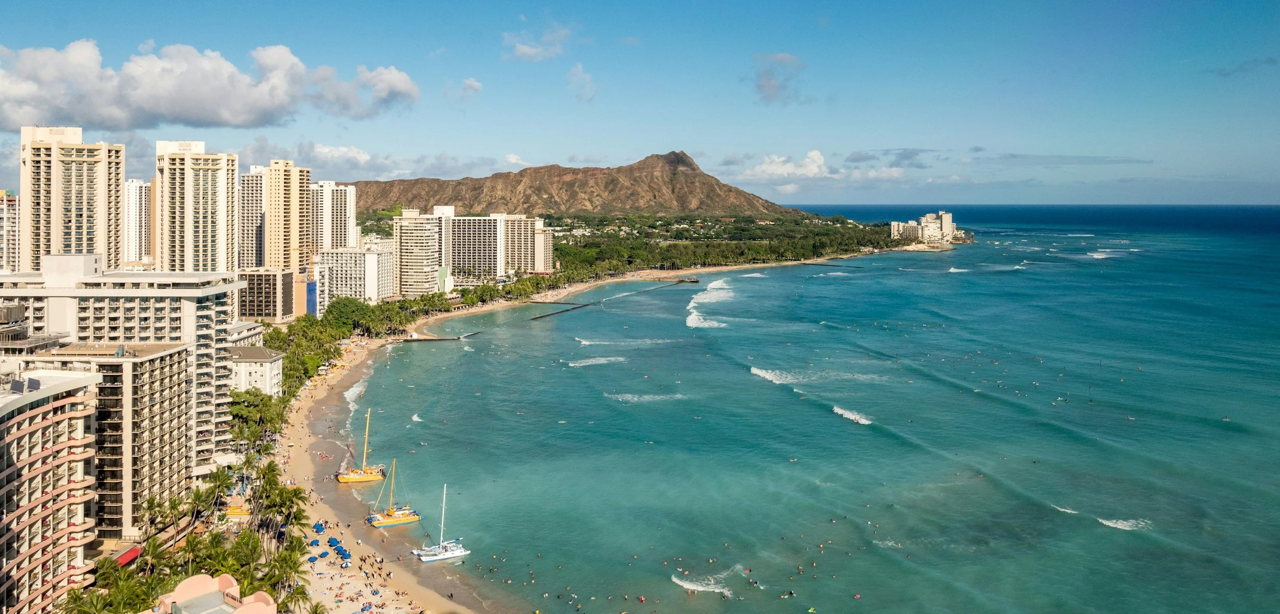 Aerial view of a tropical beach with high-rise hotels, palm trees, boats on the water, and a mountain in the background under a partly cloudy sky.