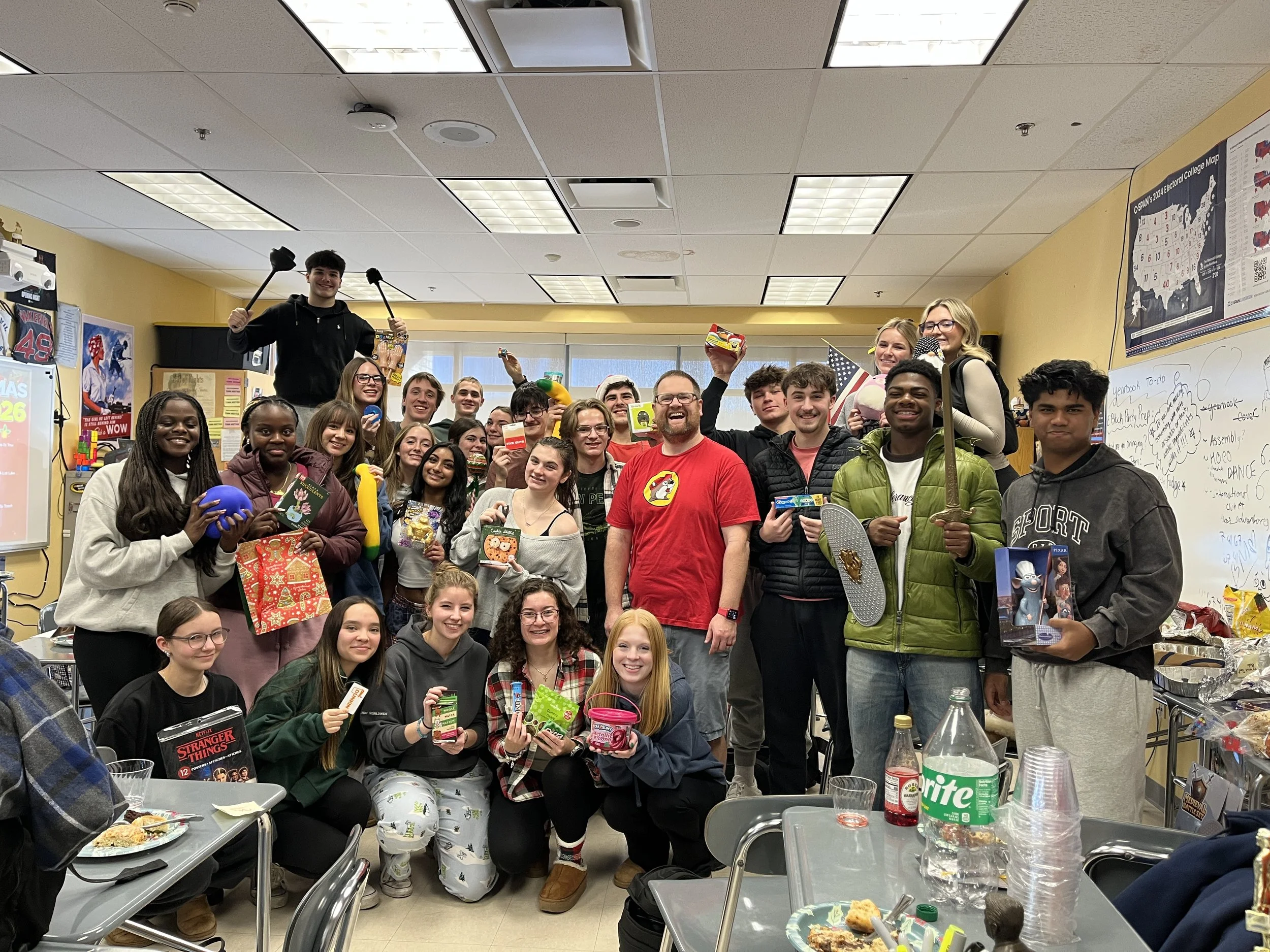 Group of students and teacher celebrating in a classroom, holding snacks, toys, and gifts, with a whiteboard and classroom decorations in the background.