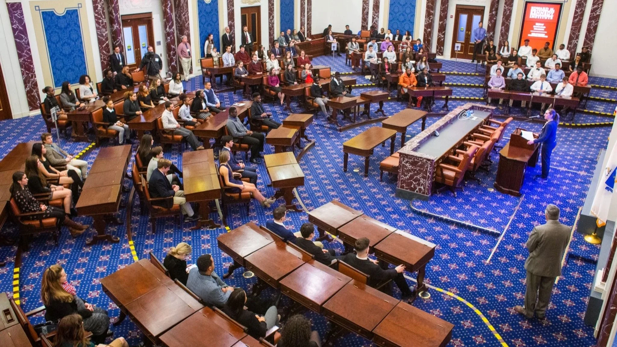 A crowd of people seated in a large, formal legislative chamber replicated after the U.S. Senate Chamber, listening to a woman speaking at a podium on the right side of the image.