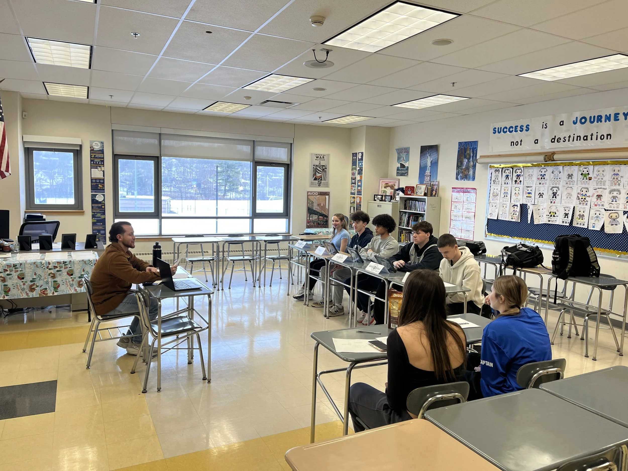 Teacher sitting at a desk with a laptop in front of students seated at desks in a classroom with large windows, posters, and decorations on the walls.