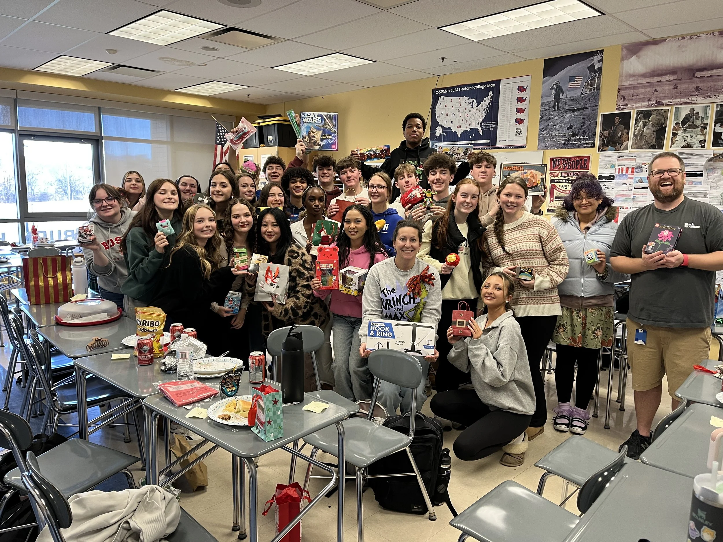 Group of students and a teacher in a classroom celebrating a holiday, holding gifts and party treats, with tables and classroom decorations visible.