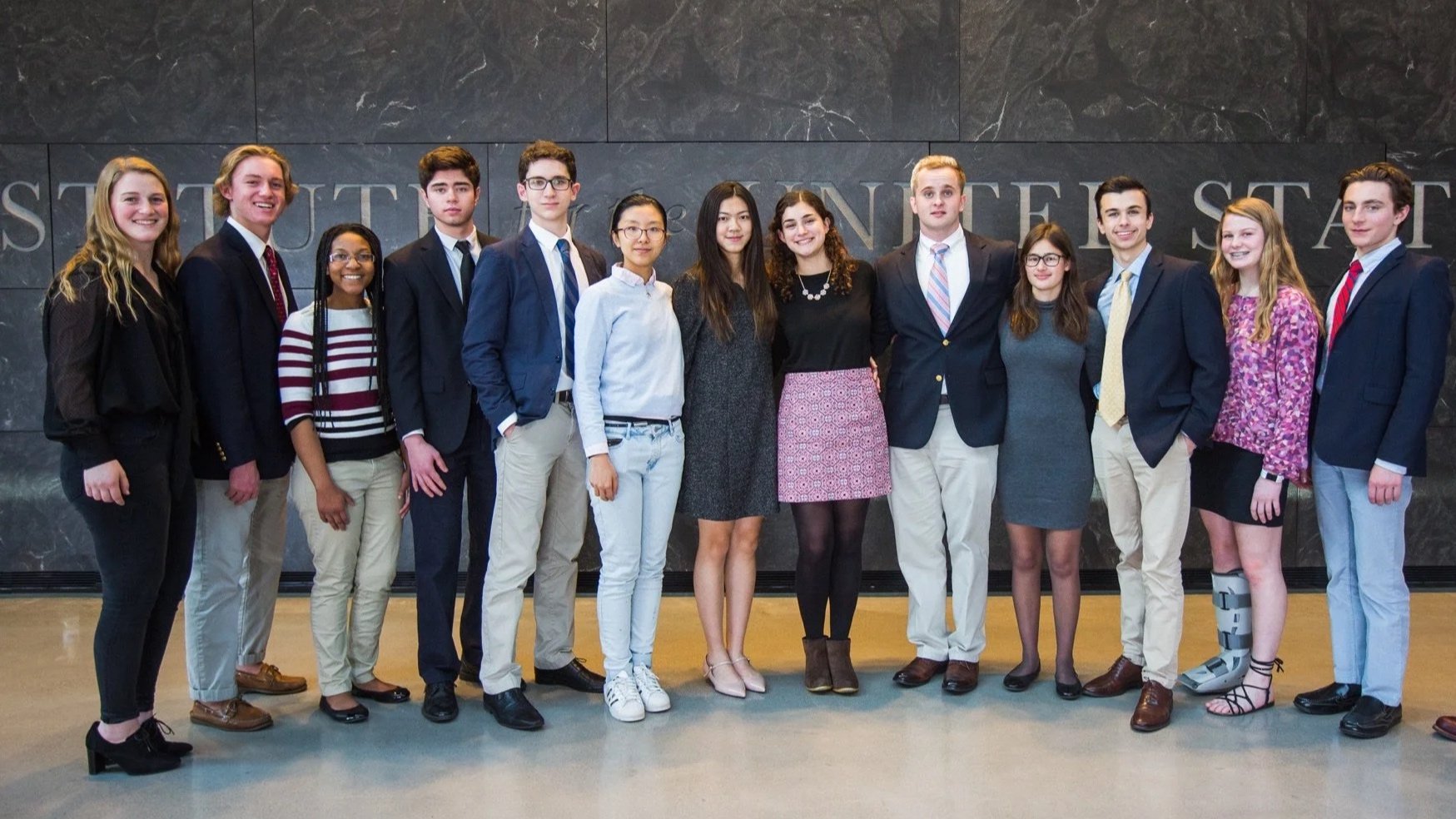 Group of young adults standing in a line indoors with a stone wall background.