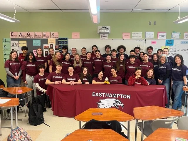 Group of students and teachers in a classroom, with a table that says 'EASTHAMPTON' and features a hawk logo.