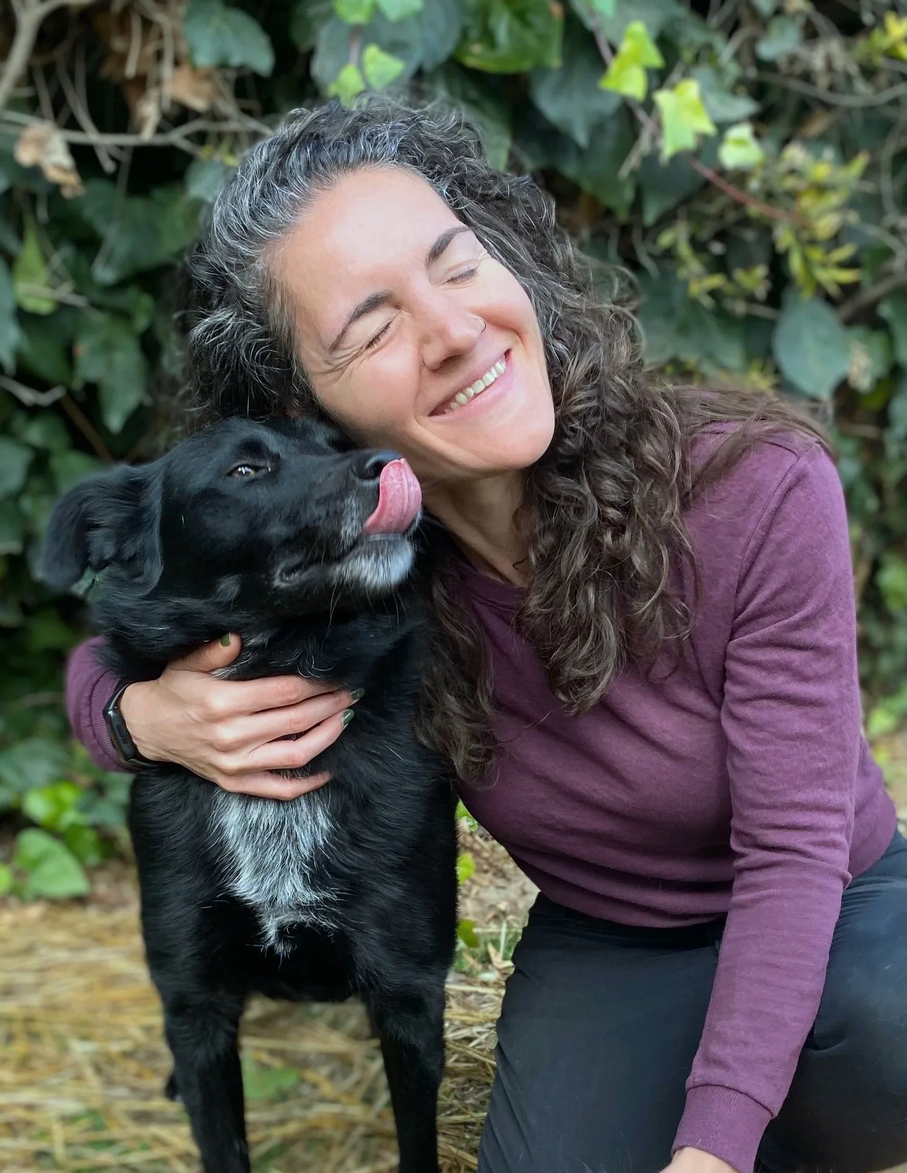 A woman with curly hair smiling with her eyes closed, hugging a black dog with white markings, surrounded by green foliage.