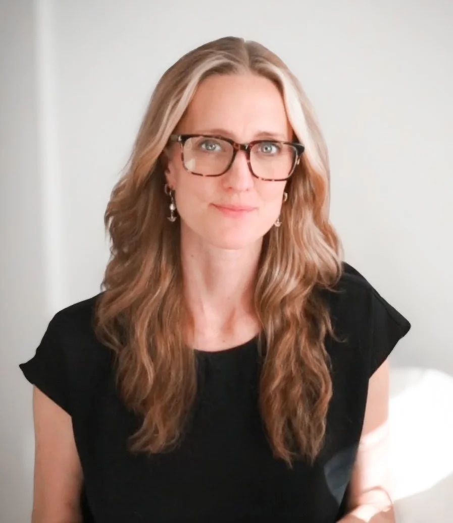 A woman with long wavy blonde hair, wearing glasses and earrings, in a black top, standing indoors in front of a plain white wall.