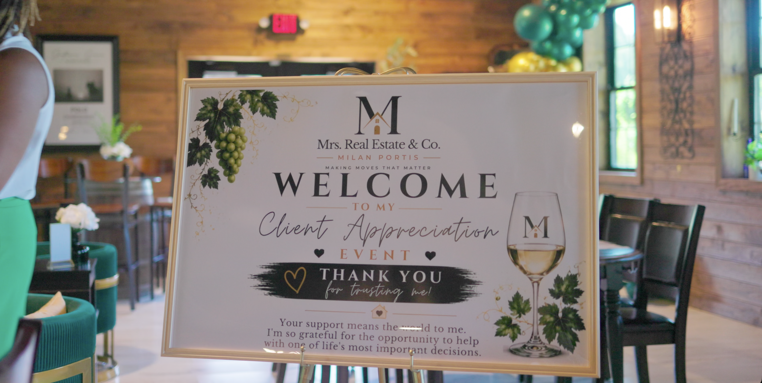 Event welcome sign with the Mrs. Real Estate & Co. logo, floral design, and a glass of white wine, acknowledging client appreciation, inside a rustic-style venue with wooden walls and balloon decorations.