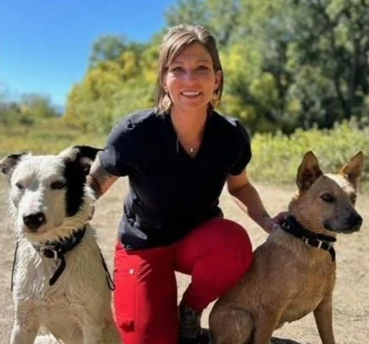 A woman crouching outdoors on a sunny day, smiling, with two dogs on either side of her. The dog on the left is a black and white Border Collie, and the dog on the right is a brown mixed breed. Green trees and a clear blue sky are in the background.