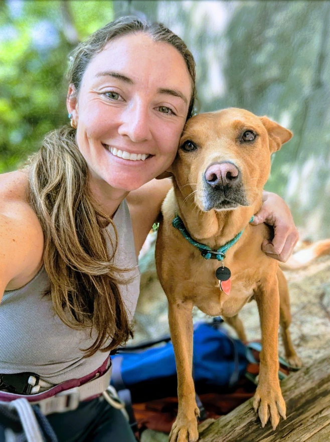 A woman who works at Mountain View Veterinary Hospital, with long brown hair and a gray tank top, smiling, taking a selfie with a brown dog with a blue collar in a forest setting.