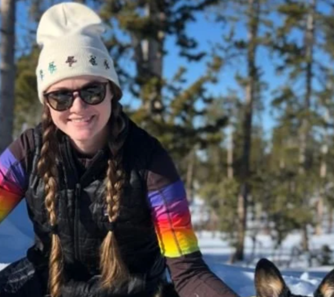 A woman at  Mountain View Veterinary Hospital, with long braided hair, wearing sunglasses, a white beanie with colorful designs, and a rainbow-colored sleeve winter jacket, smiling outdoors in a snowy forest setting.