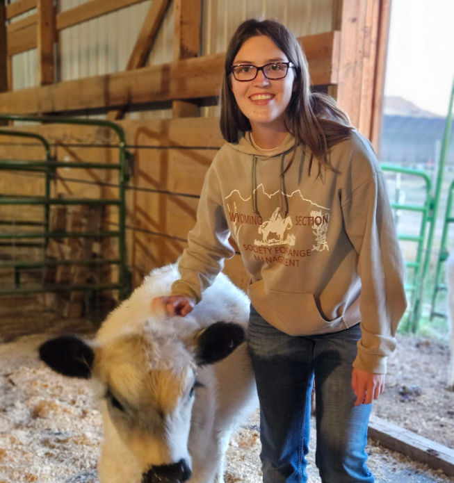 Young woman at  Mountain View Veterinary Hospital, with glasses and a beige hoodie smiling while petting a small white calf in a barn.