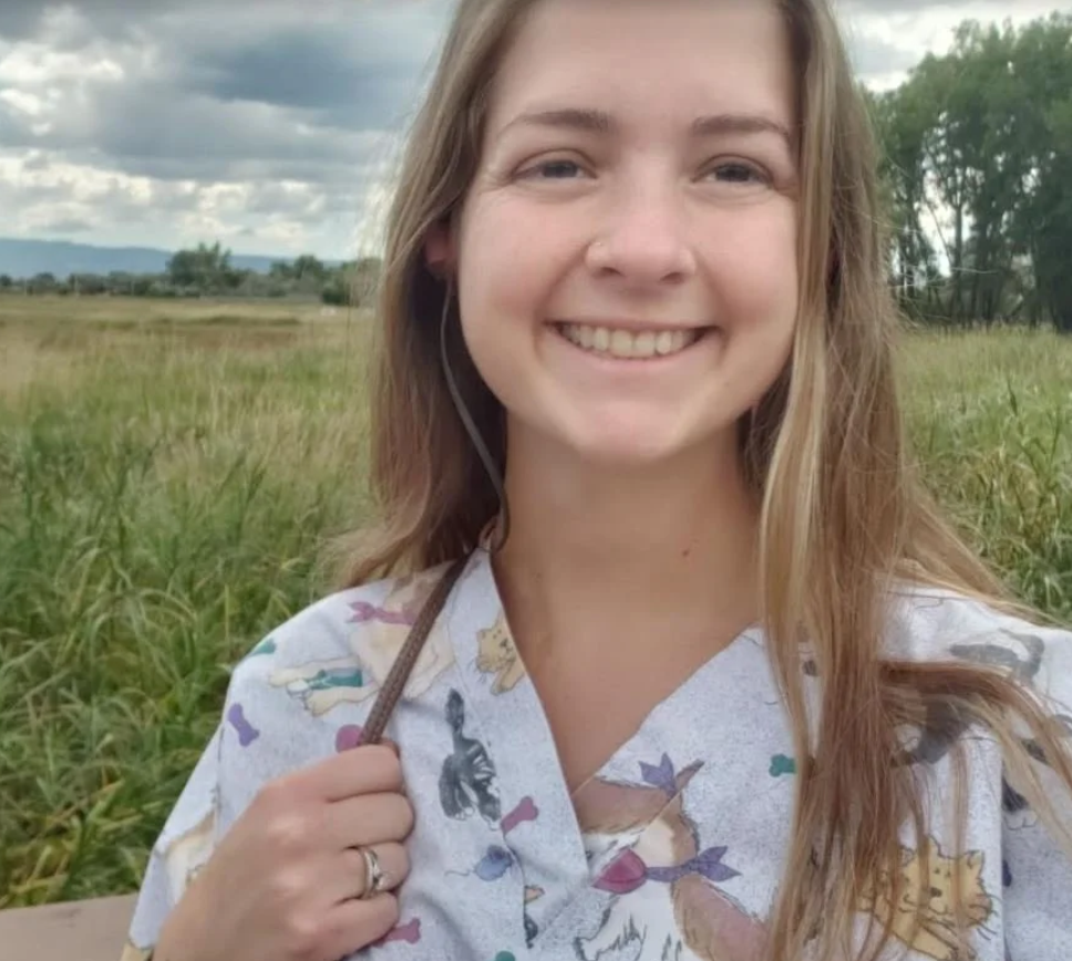 A smiling young woman at  Mountain View Veterinary Hospital, with long hair standing outdoors in a grassy field with trees and cloudy sky in the background, wearing a patterned shirt and a backpack strap over her shoulder.