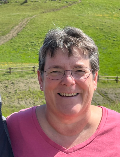A smiling woman at Mountain View Veterinary Hospital,with short gray hair and glasses, wearing a pink shirt, outdoors on a sunny day with green hills in the background.