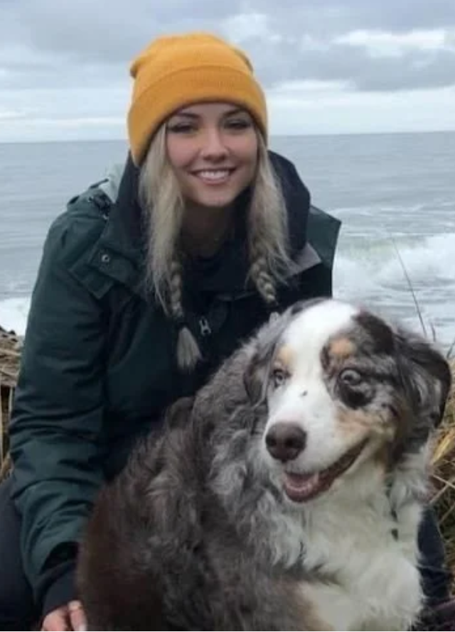A young woman at Mountain View Veterinary Hospital, with blonde hair in braids, wearing an orange beanie and dark jacket, smiling while outdoors near water with a large, fluffy Australian Shepherd dog.