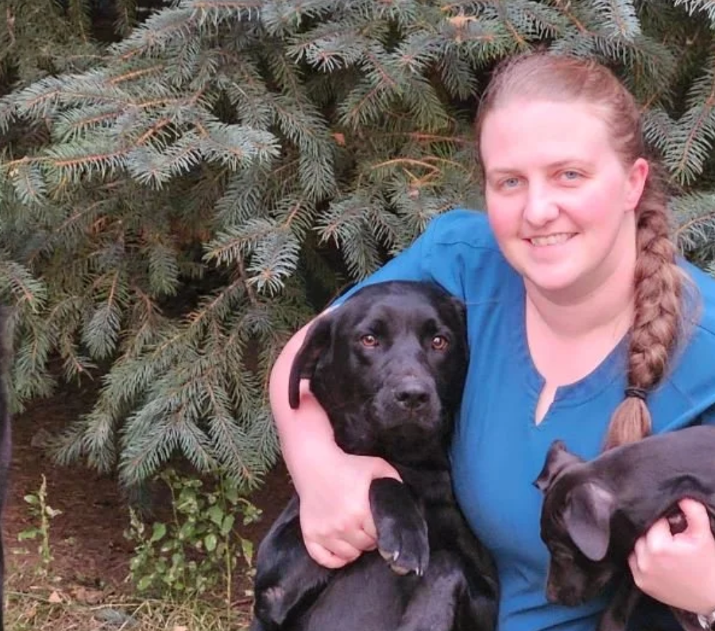 A woman at  Mountain View Veterinary Hospital, with long braided hair smiling while holding two black dogs, in front of a pine tree.