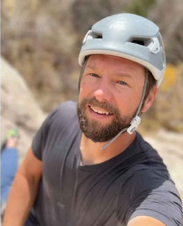 A man at  Mountain View Veterinary Hospital, with a beard wearing a bike helmet and a black t-shirt taking a selfie outdoors with a blurred background of rocks and foliage.