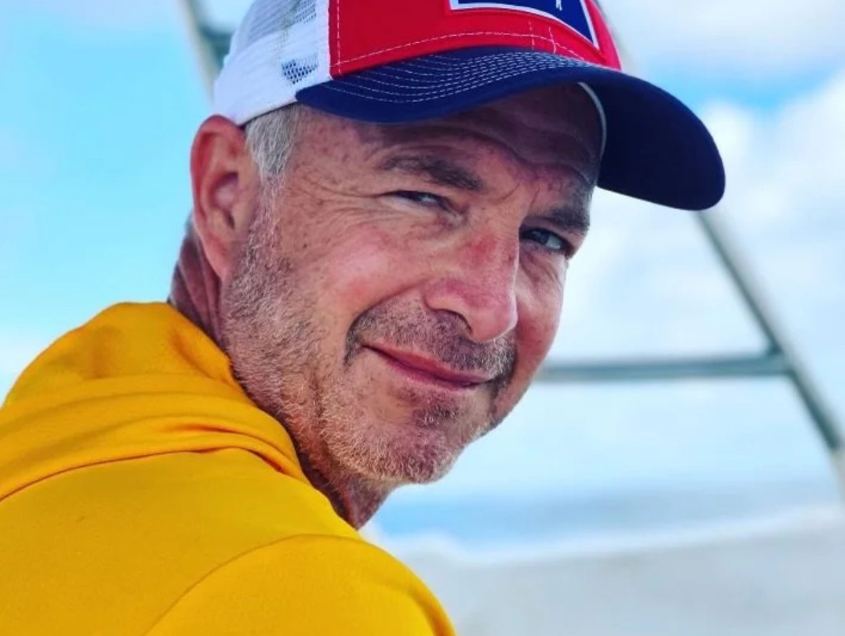 Close-up of a smiling man at  Mountain View Veterinary Hospital, with a beard wearing a yellow shirt and a red, white, and blue cap with a blue sun visor, outdoors under a cloudy sky.