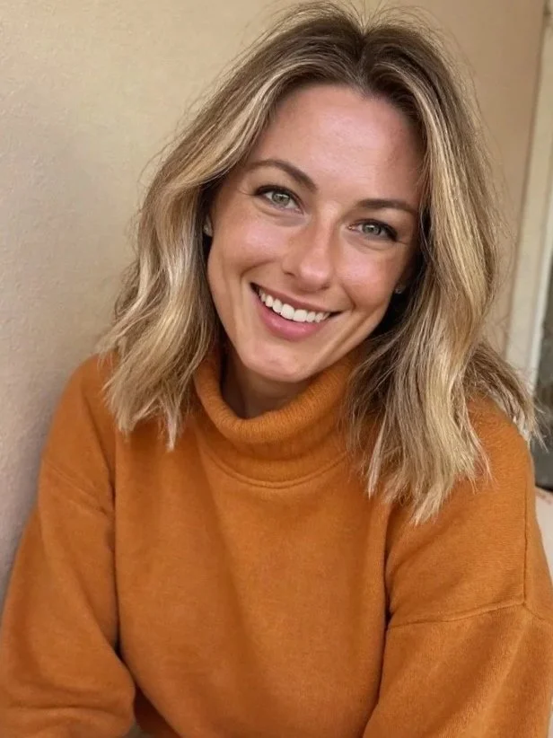 A young woman with shoulder-length blond curly hair smiling, wearing an orange turtleneck sweater, and sitting against a beige wall.