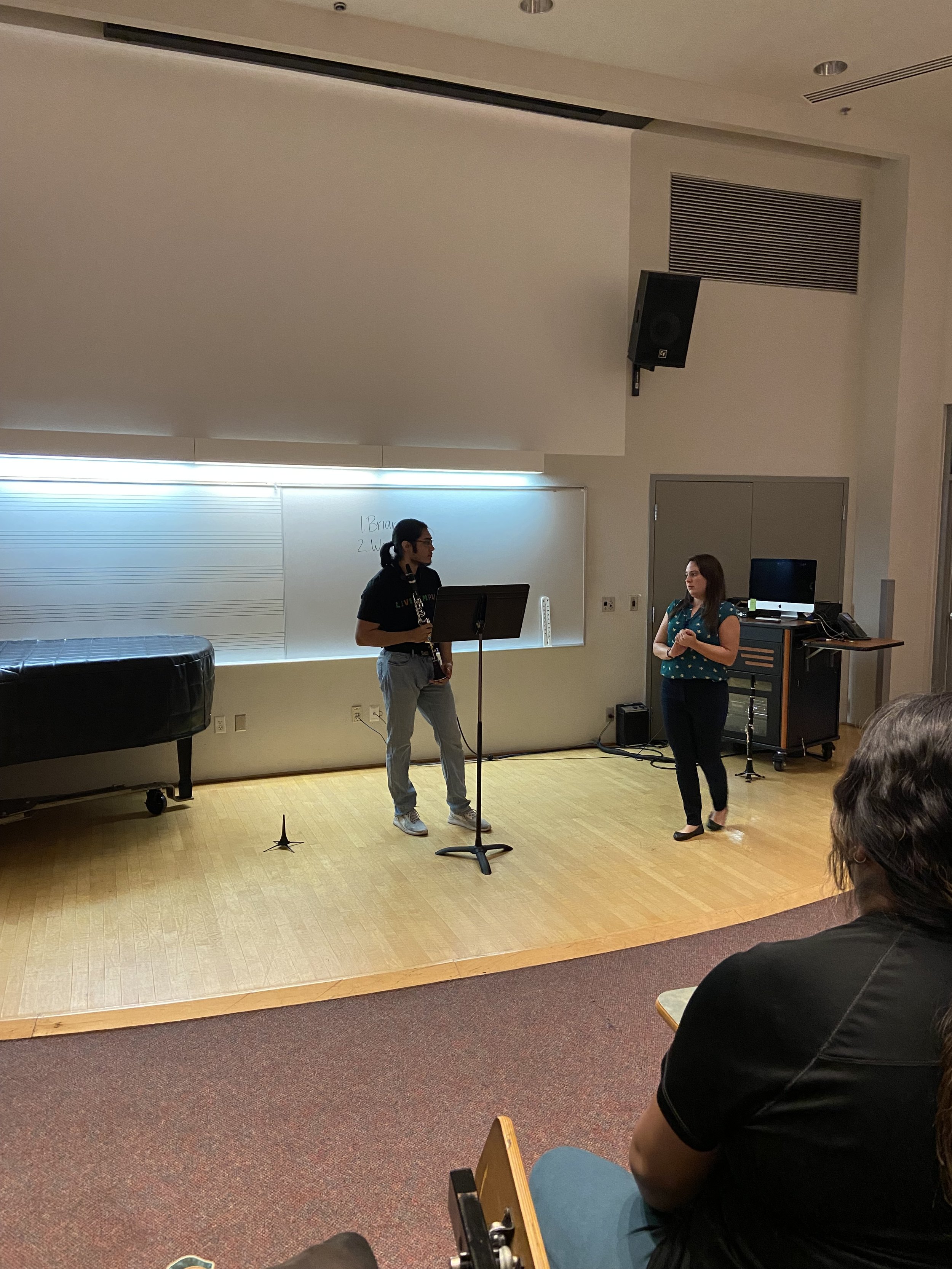 A classroom or lecture hall with two women standing at the front. One woman is holding a clarinet, and both are engaged in a discussion. There is a music staff whiteboard, a piano, and a computer setup present in the room. Audience members are seated and facing the women.