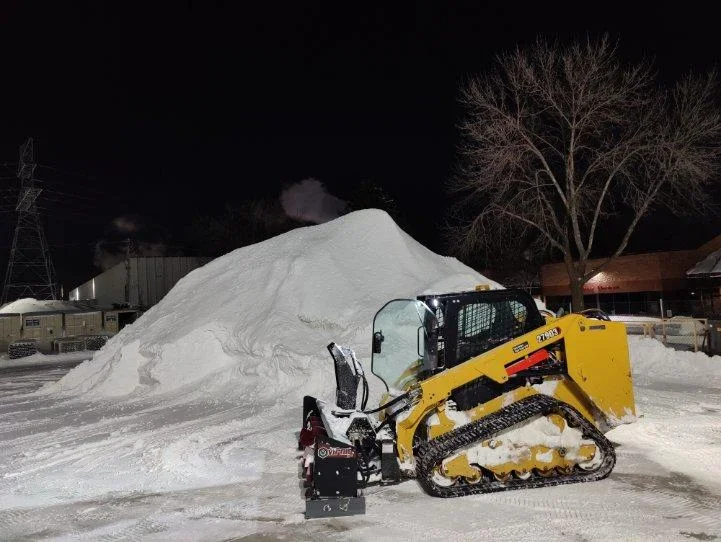 Snow removal vehicle clearing snow from a road surrounded by trees.