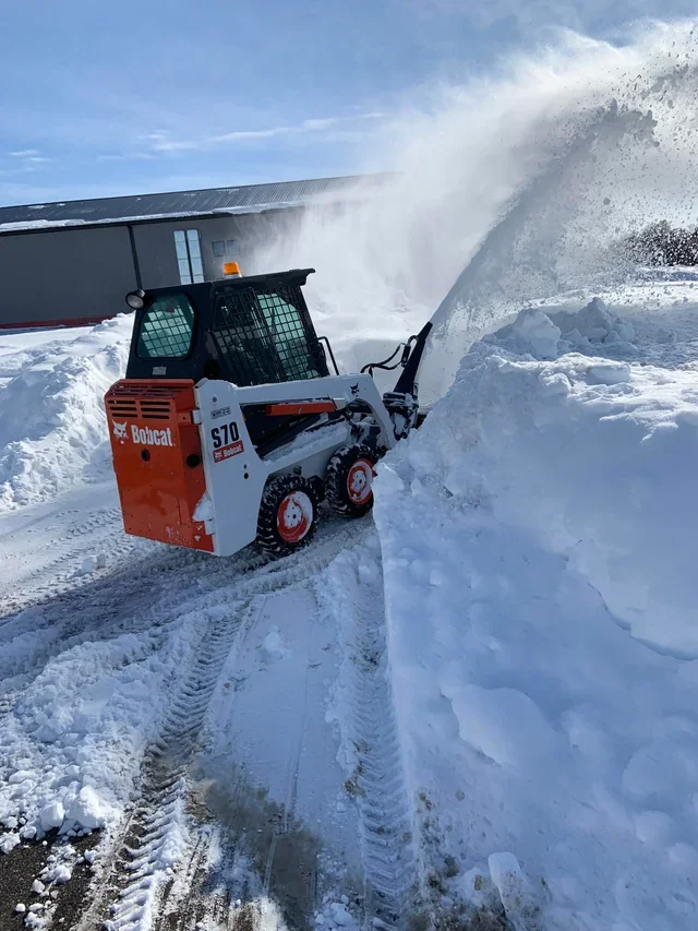 A compact Bobcat S70 skid-steer loader clearing snow with a bucket attachment in a snowy outdoor area. Snow is being pushed aside, creating a large snowbank, with a building visible in the background under a partly cloudy sky.