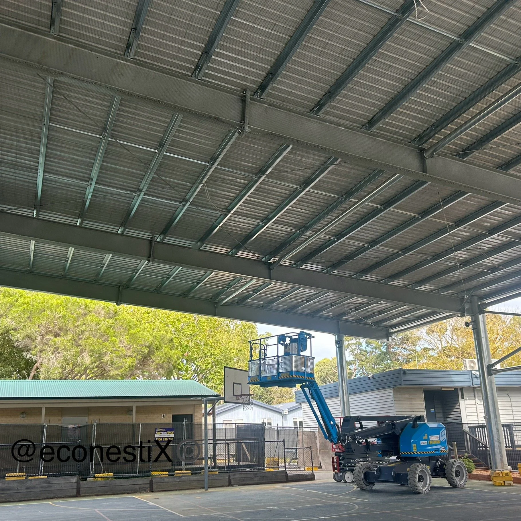 Basketball courts being netted towards the completion of the structure being built. Shows height access equipment a Boom lift on the court which was used by qualified workers to undertake work a heights along with another operator on a EWP Scissor li