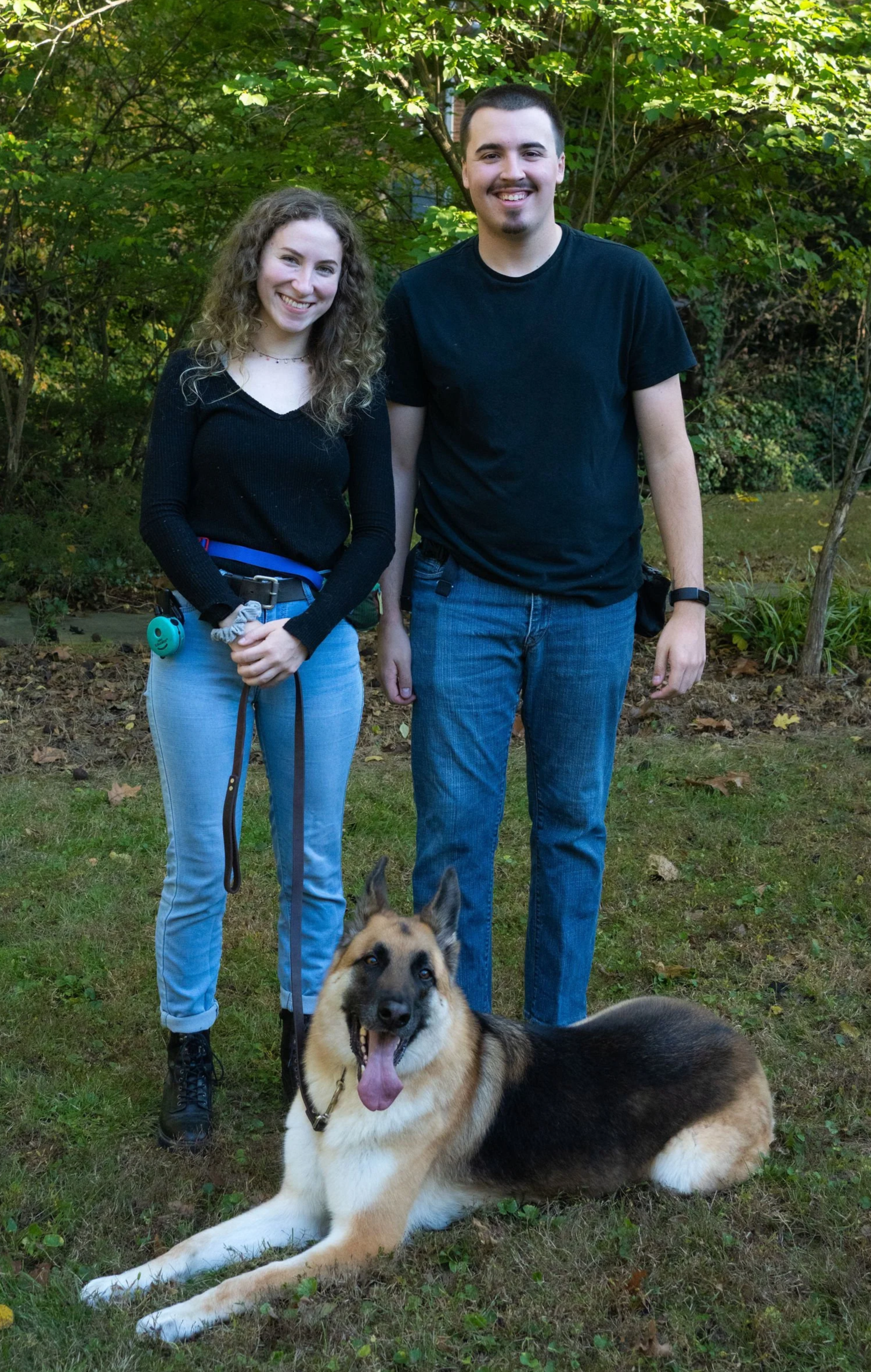 A young woman and man standing outdoors with a German Shepherd dog lying on the grass in front of them, surrounded by trees and plants.