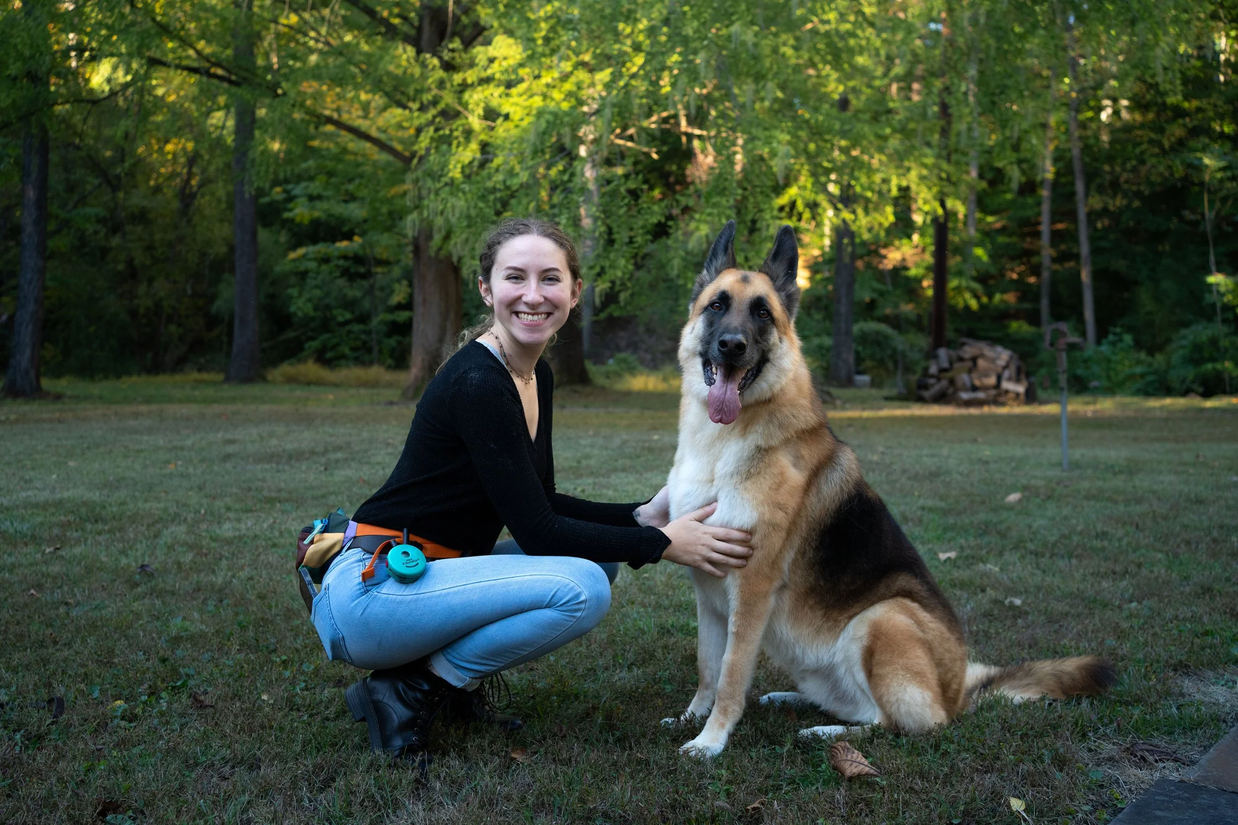 A young woman in black sweater and light blue jeans squats on grass, smiling, with her hand on a German Shepherd dog sitting next to her in a park with green trees in the background.