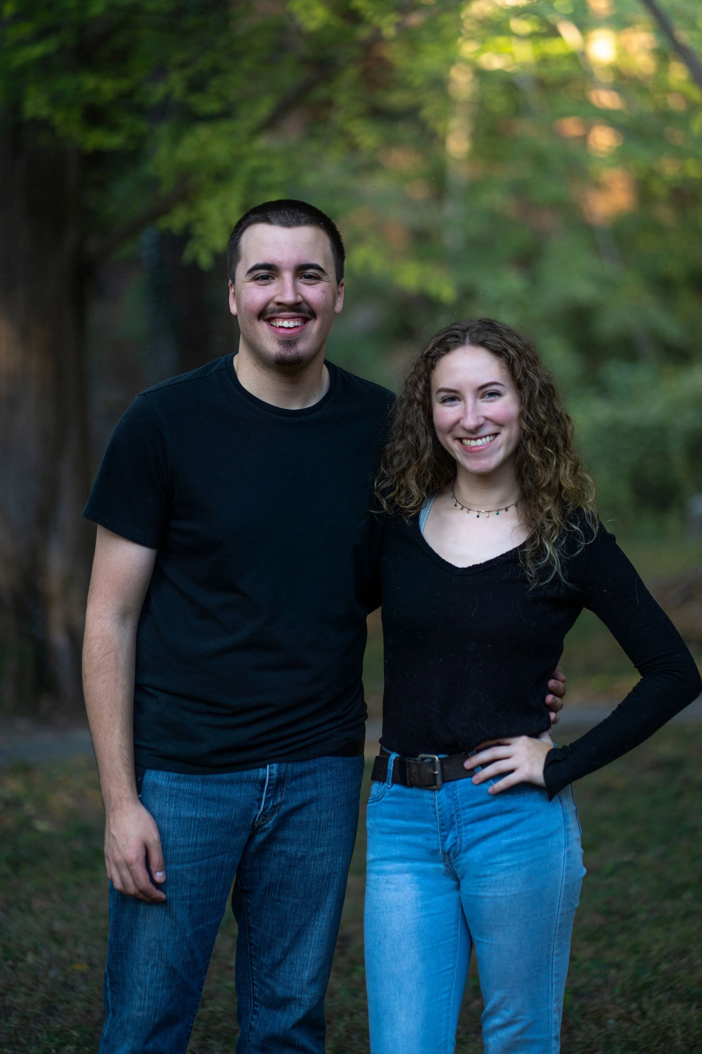 A young man and woman standing together outdoors in a park, smiling at the camera with trees and greenery in the background.