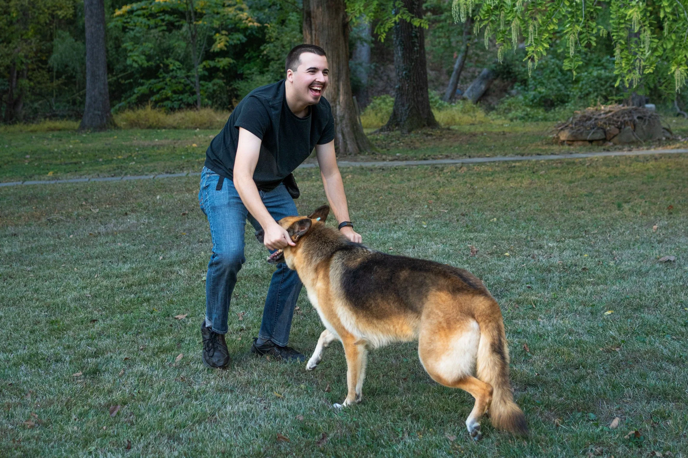 A man playing with a German Shepherd dog in a grassy park area with trees in the background, smiling and laughing.