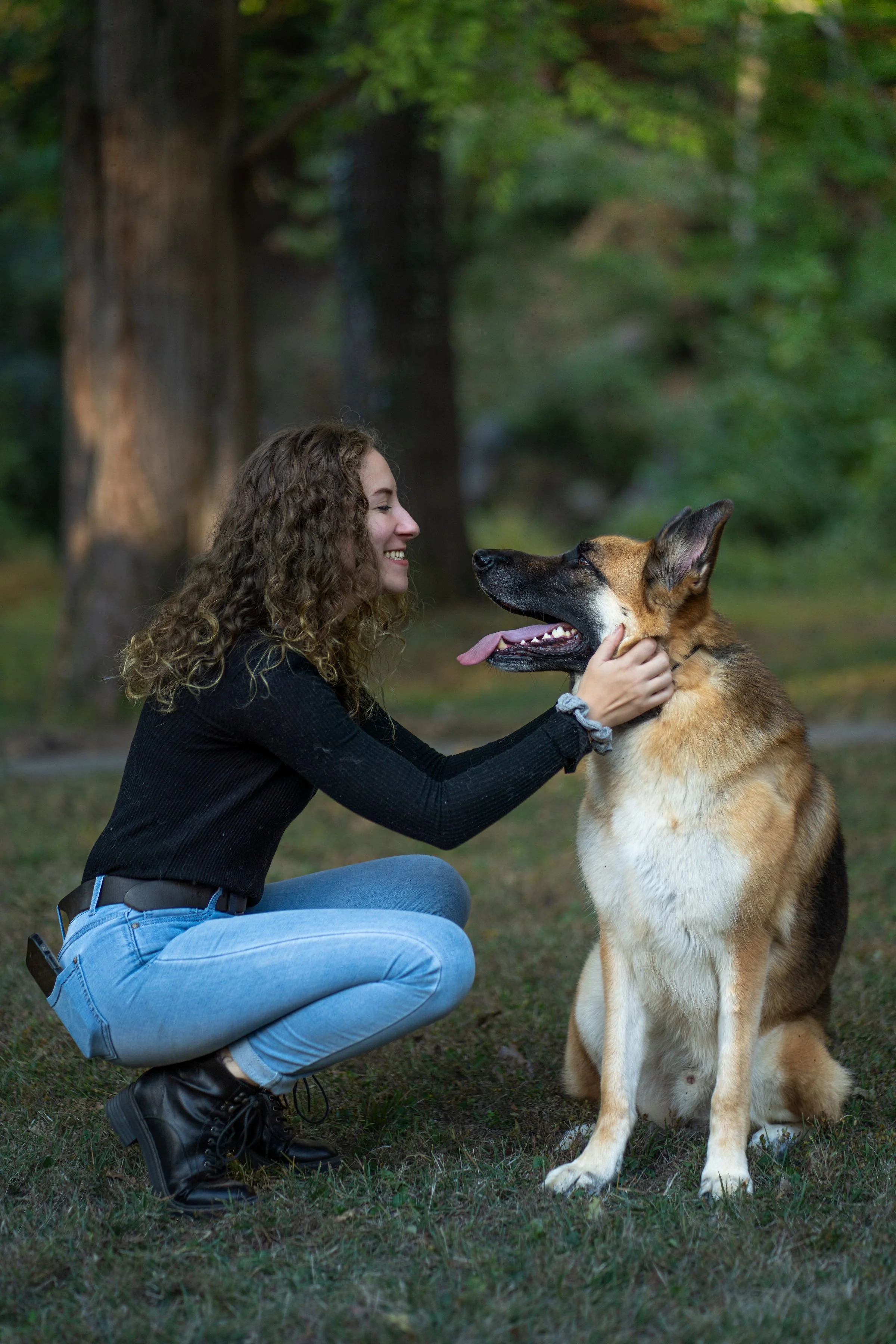 A woman with curly hair crouches down and smiles at a large German Shepherd dog outdoors in a park with tall trees.