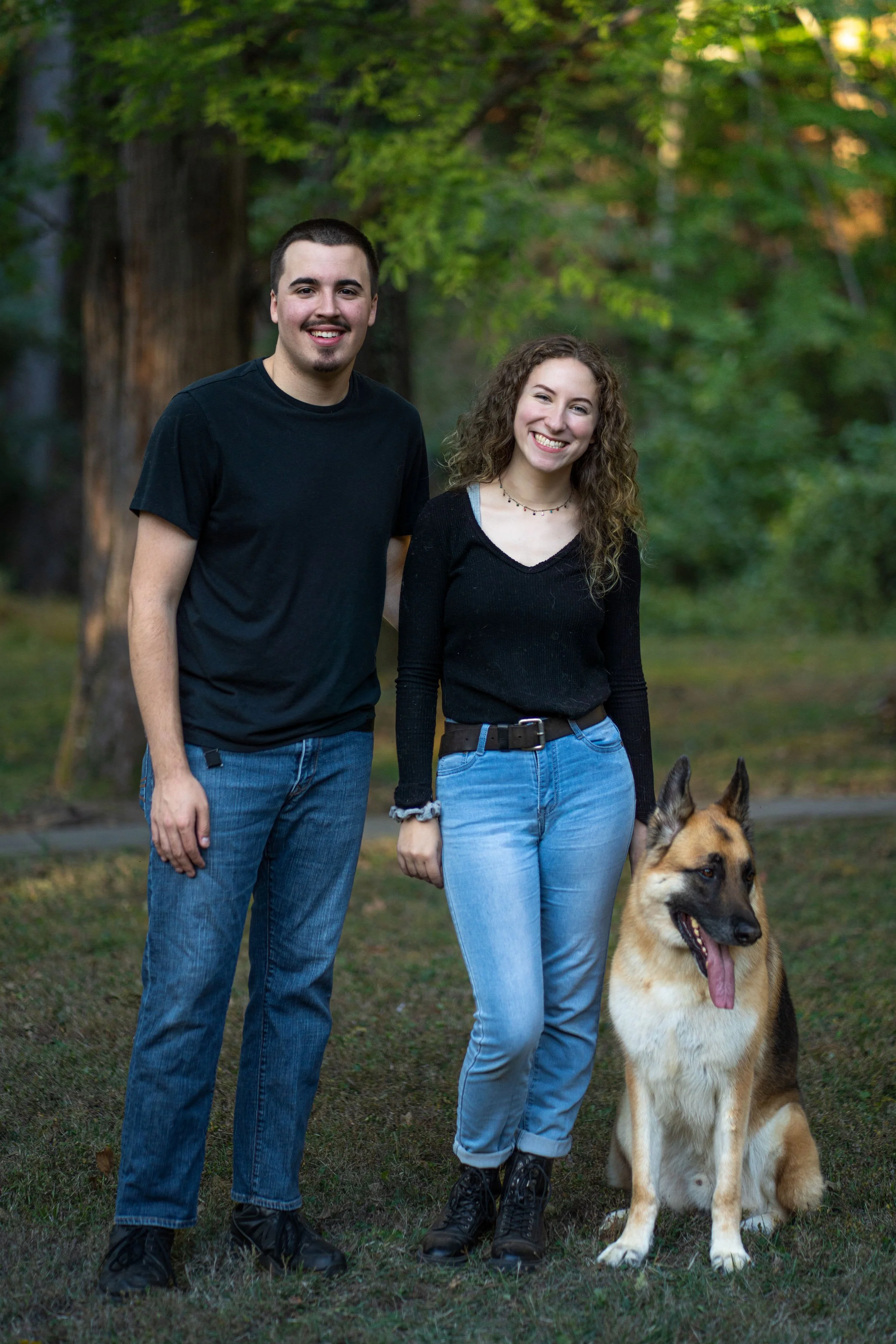 A young man and woman standing outdoors with a German Shepherd dog, trees in the background, smiling for the camera.