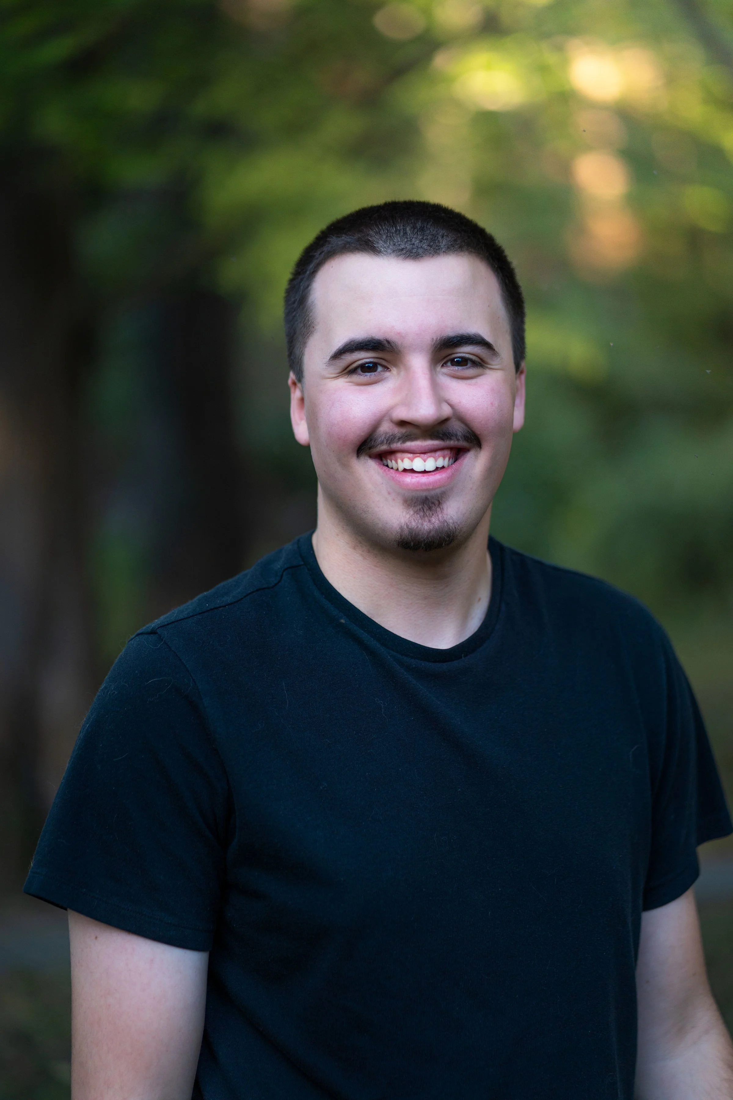 A young man with short dark hair and a goatee smiling outdoors, wearing a black t-shirt with a blurred green and yellow background.