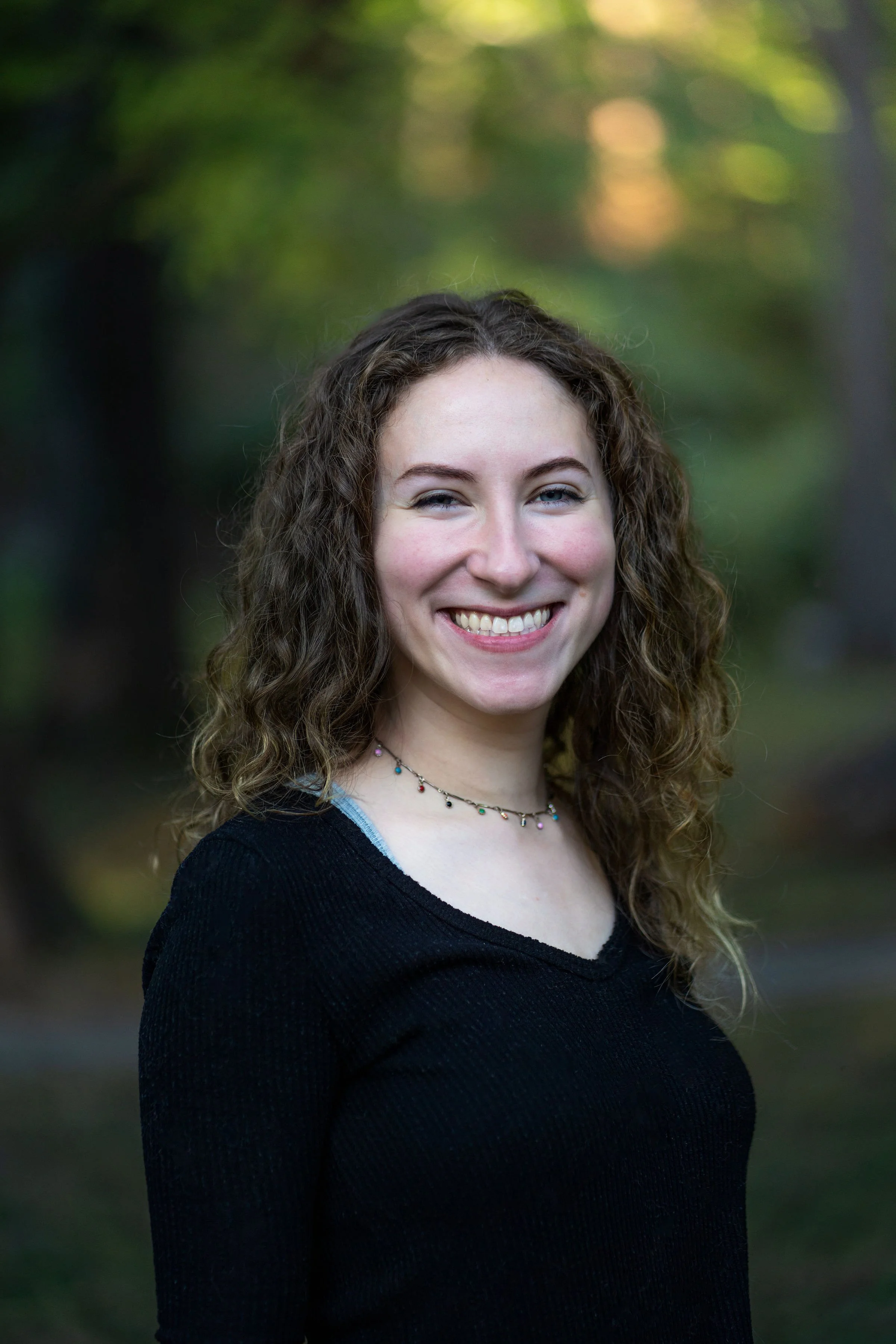 A young woman with curly hair smiling outdoors, wearing a black top and colorful beaded necklace, with trees and greenery in the background.