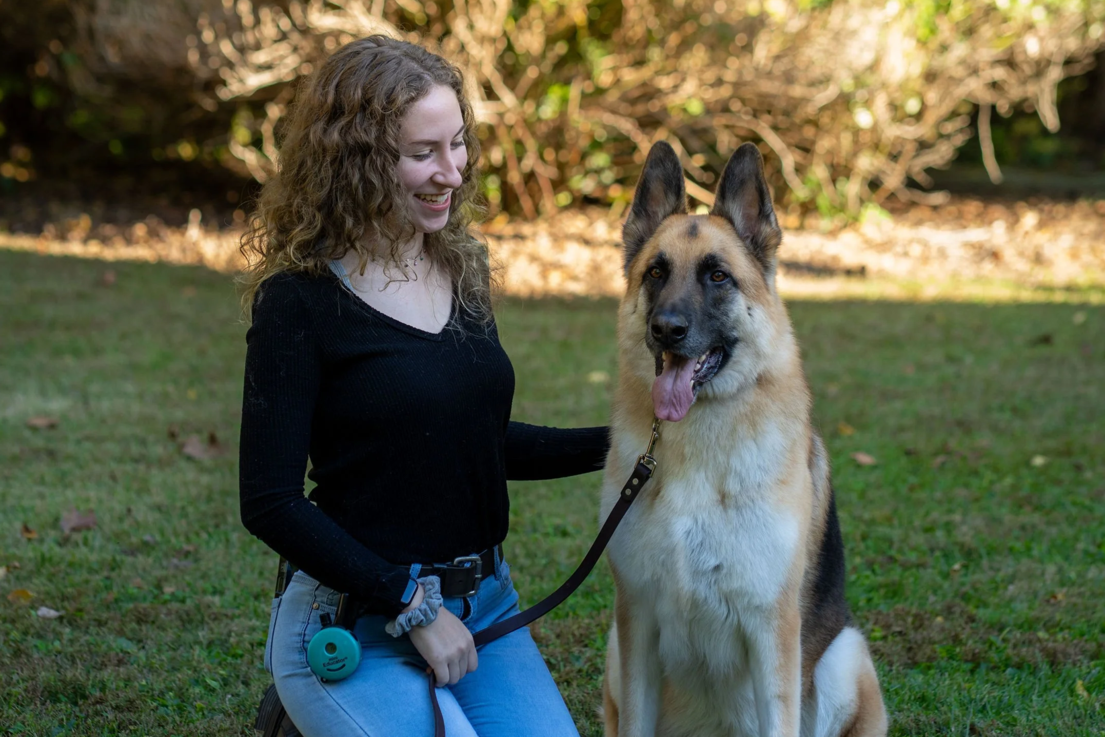 A woman smiling and sitting on the grass outdoors with an excited German Shepherd dog on a leash.