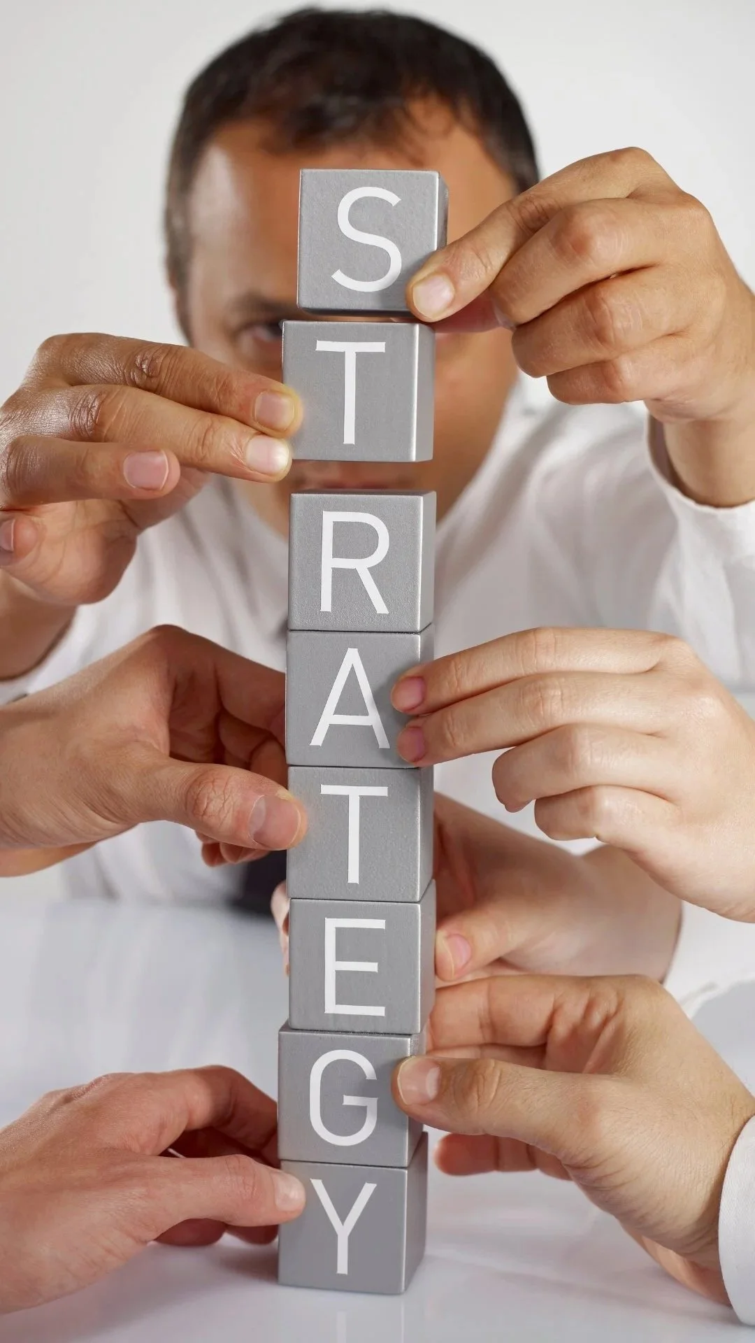 Multiple hands stack gray blocks vertically, spelling out the word 'STRATEGY' in white letters, with a man peering behind them.