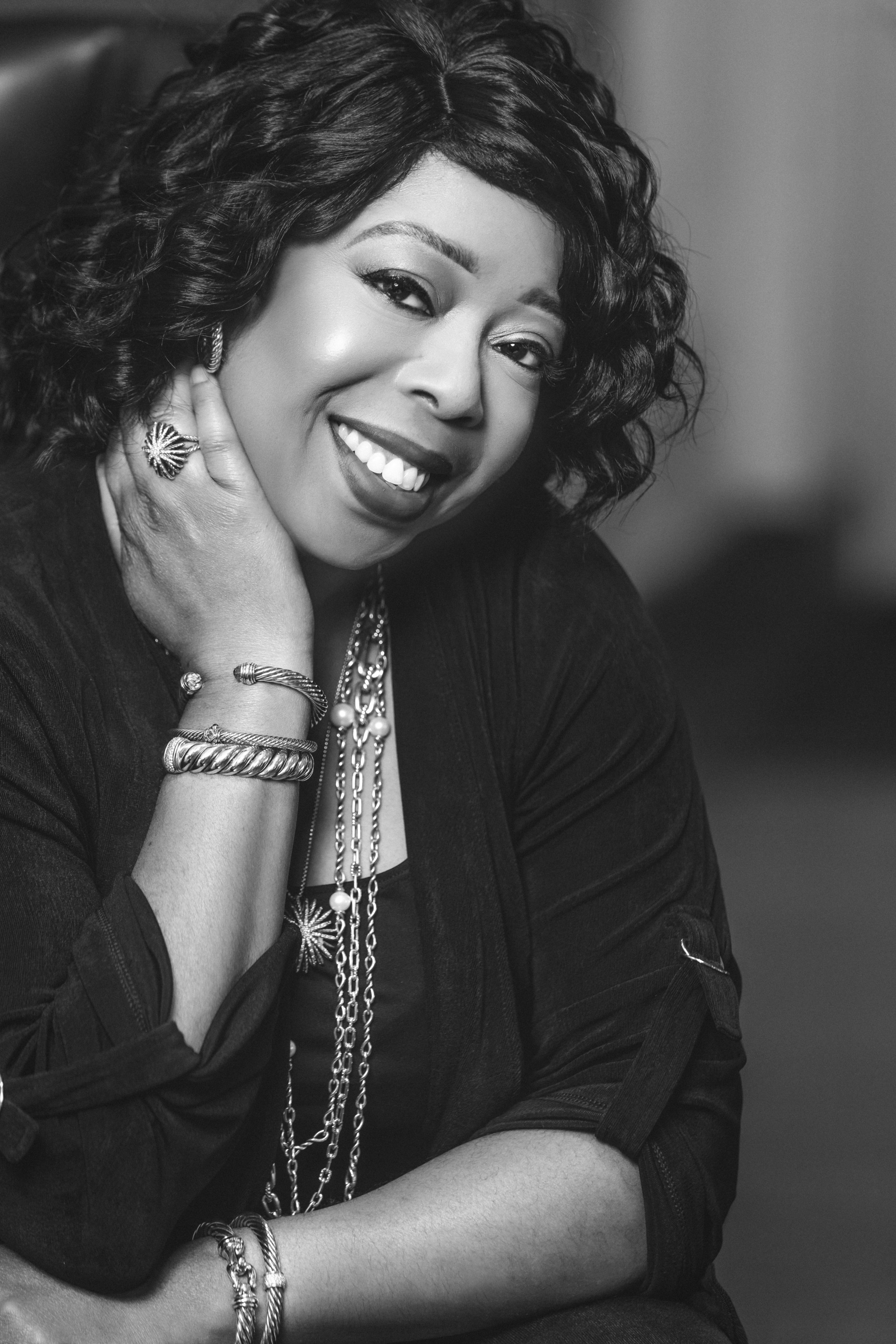 Black and white portrait of a woman smiling, wearing jewelry including rings, bracelets, and layered necklaces, with her hand resting on her neck.