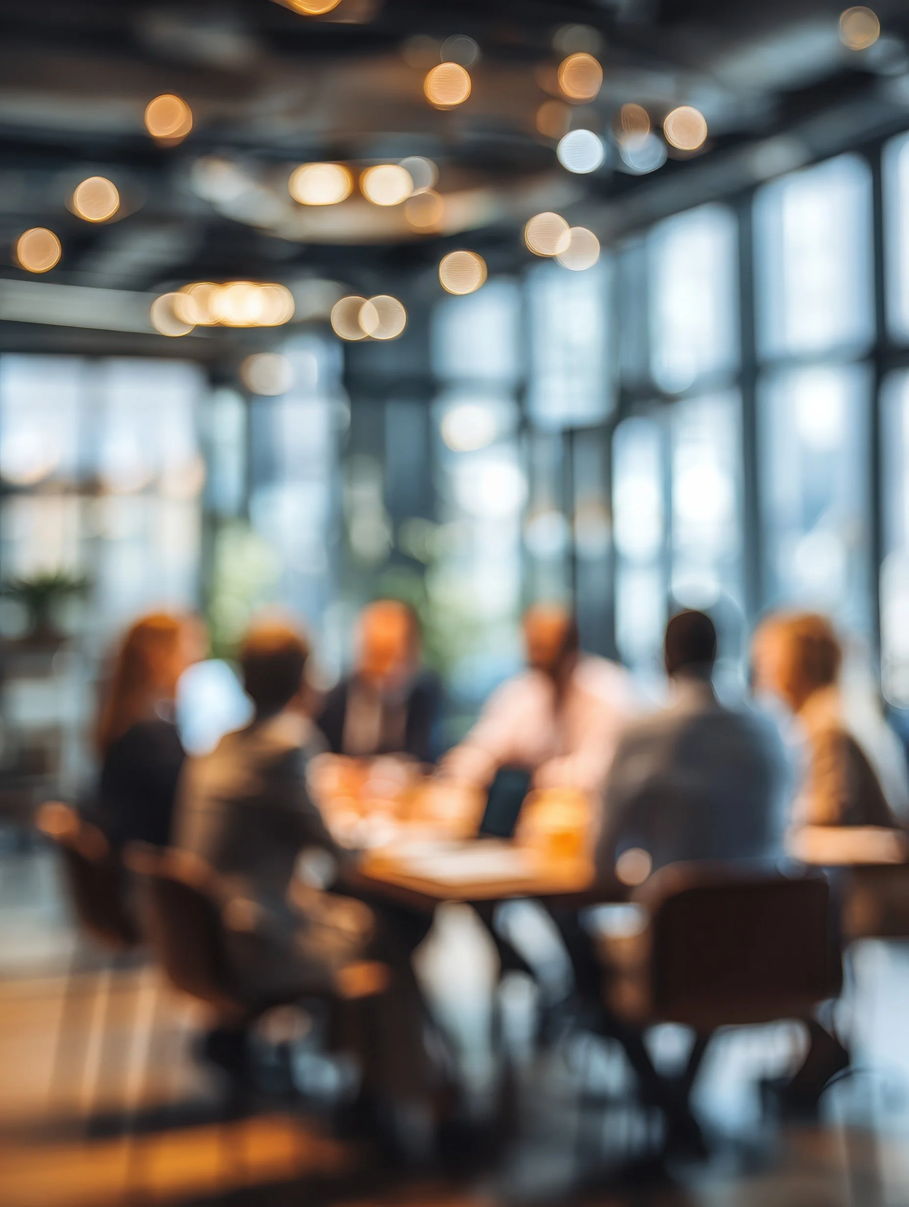 A blurry image of a group of people sitting around a table in a modern, well-lit office or conference room, with large windows and ceiling lights.