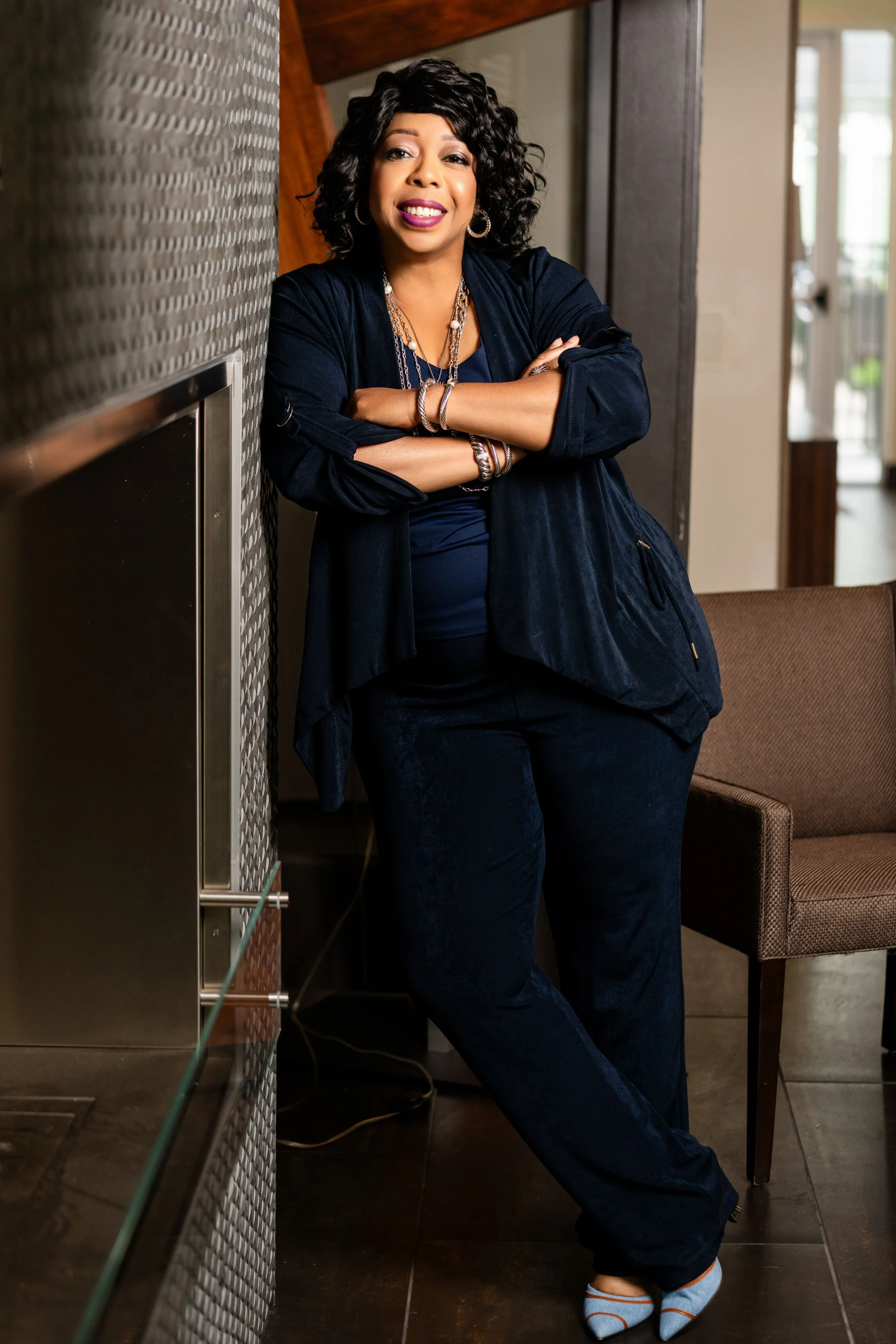 A woman with curly black hair, wearing a navy blue outfit with jewelry, leaning against a wall in an indoor setting.
