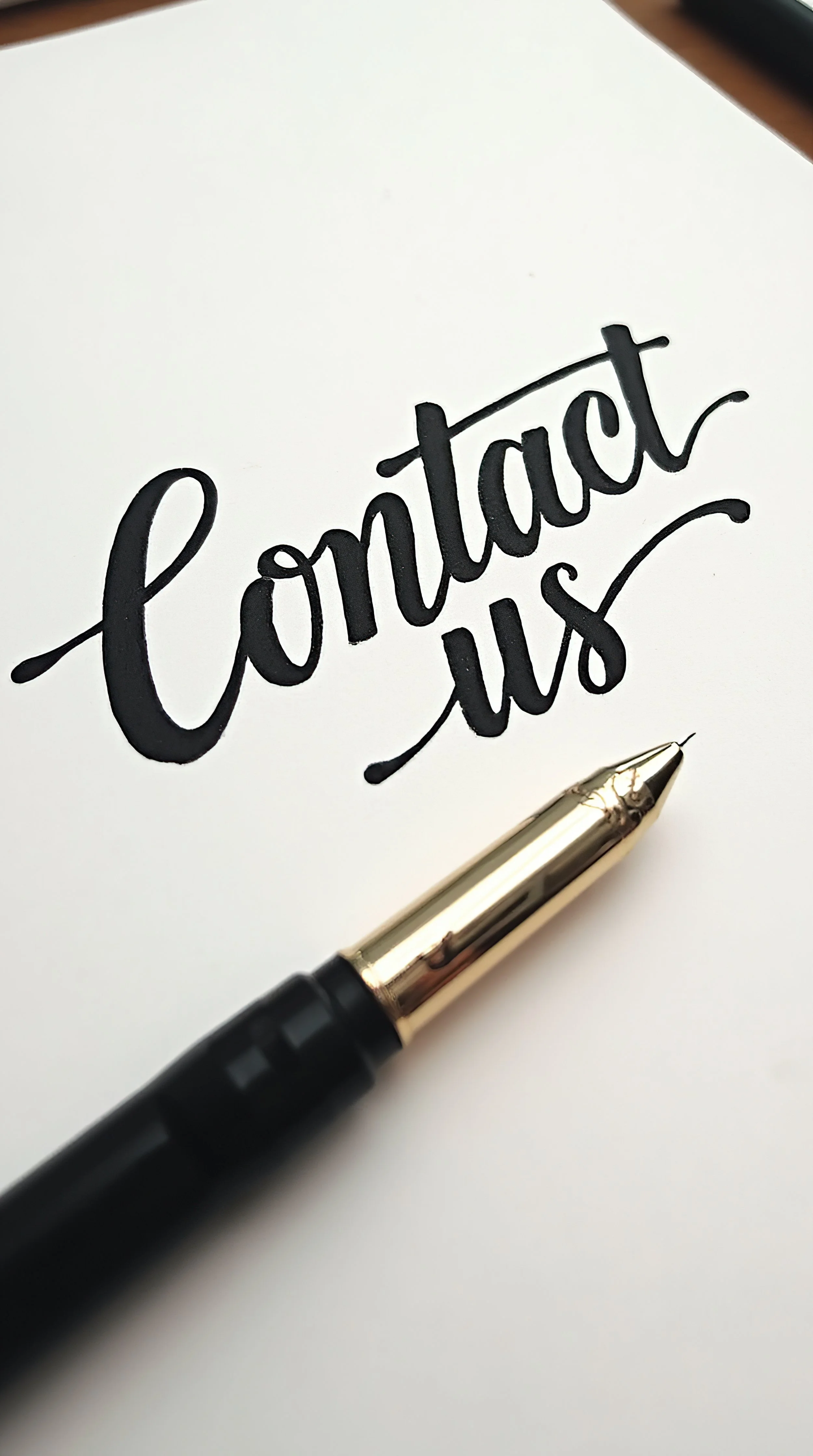 Handwritten text on white paper that says 'contact us' in black ink, with a black and gold pen resting on the paper.