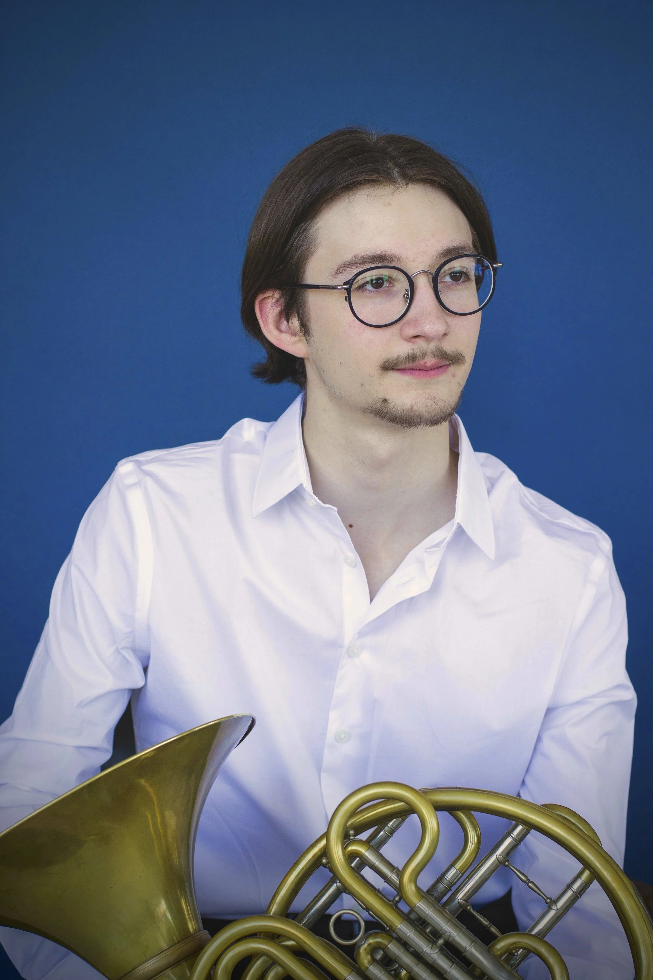 Young man with glasses and facial hair wearing a white shirt, sitting against a blue background, holding a French horn.