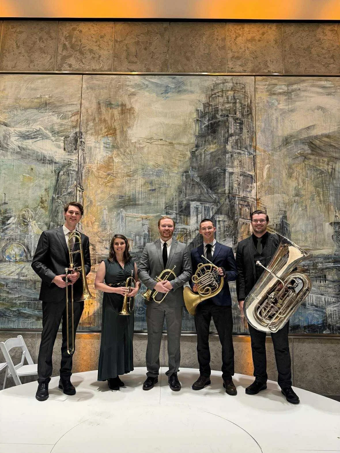 Five musicians dressed in formal attire standing in front of an abstract cityscape painting, holding brass instruments including a trombone, trumpet, French horn, and tuba.