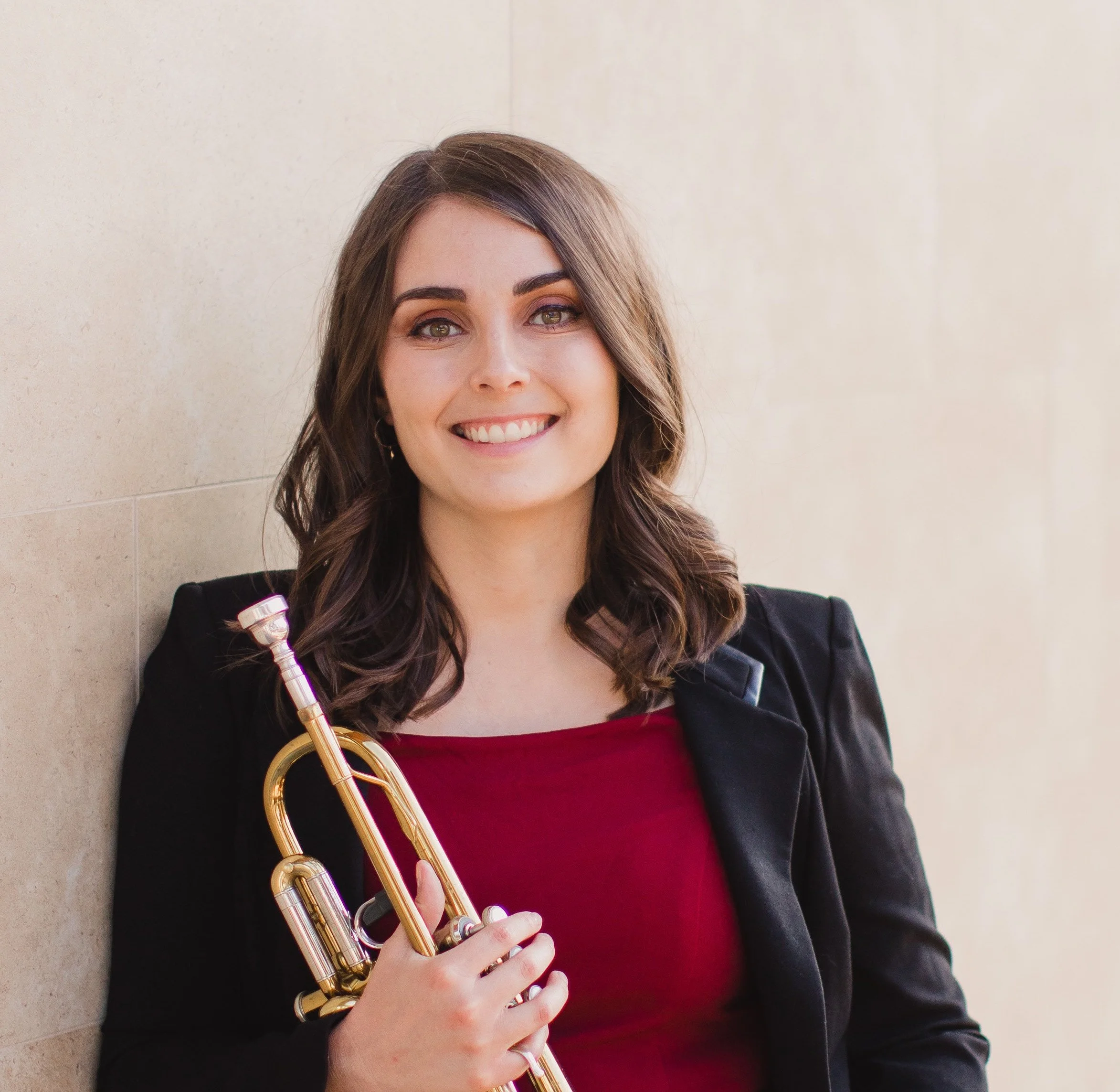 A young woman with shoulder-length brown hair, smiling, holding a trumpet, wearing a red top and black jacket, standing against a beige wall.